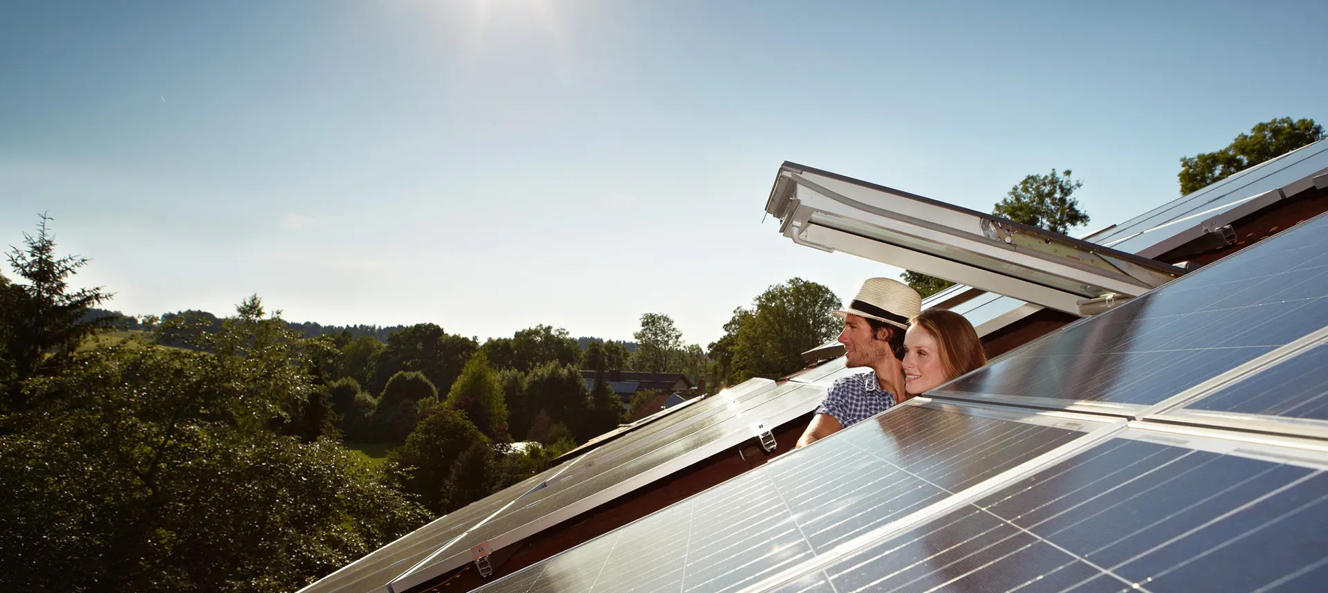 Mann und Frau schauen aus einem Dachfenster in die Ferne Mann und Frau schauen aus einem geöffneten Dachfenster. Eine PV-Anlage der NEW Energie ist zu sehen.