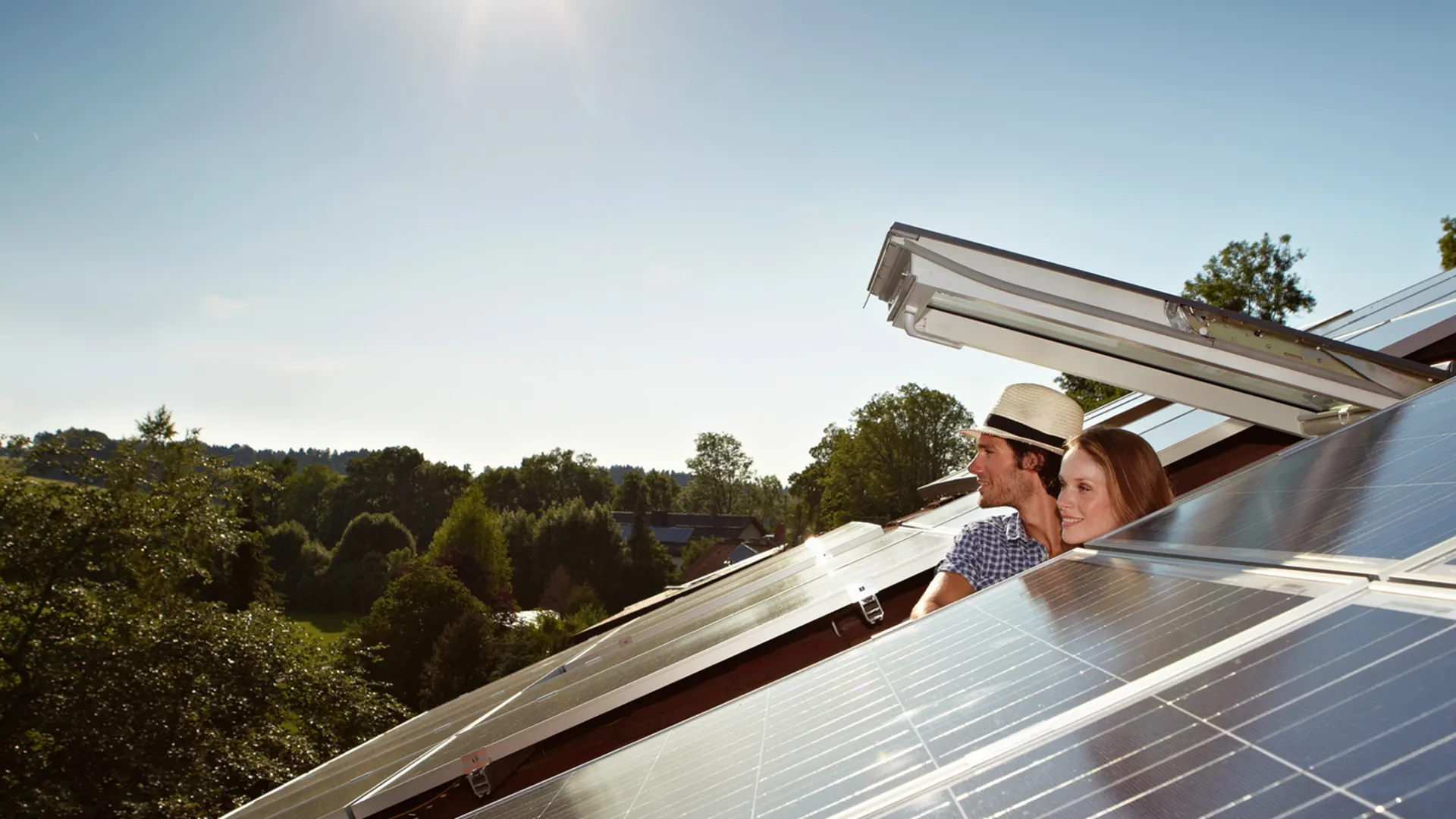 Mann und Frau schauen aus einem Dachfenster in die Ferne Mann und Frau schauen aus einem geöffneten Dachfenster. Eine PV-Anlage der NEW Energie ist zu sehen.