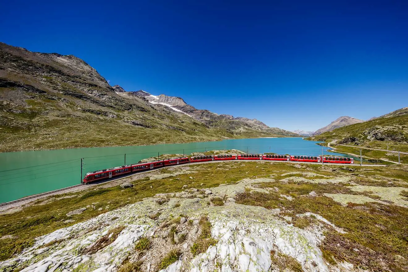 Der Bernina Express fährt entlang des türkisblauen Lago Bianco auf der Berninalinie, umgeben von Bergwelt und blauem Himmel.