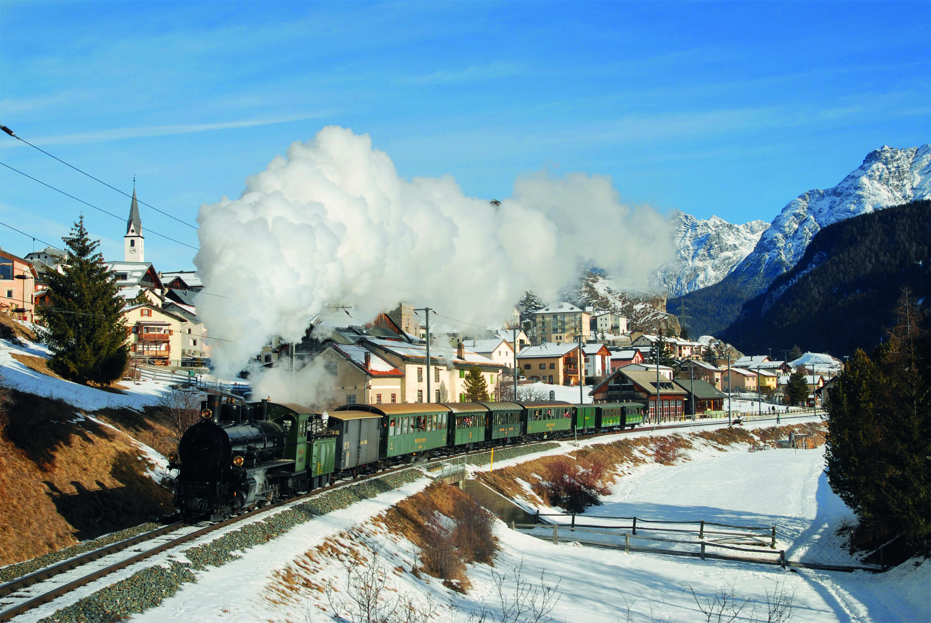 Historischer Dampfzug der RhB fährt durch das winterliche Engadiner Dorf Ardez, im Hintergrund die Gipfel des Unterengadins.