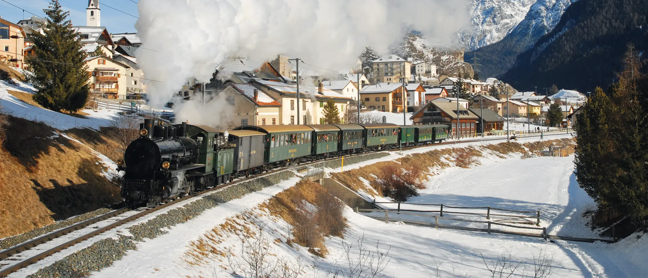 Lo storico treno a vapore della FR attraversa il villaggio engadinese di Ardez, sullo sfondo le vette della Bassa Engadina.