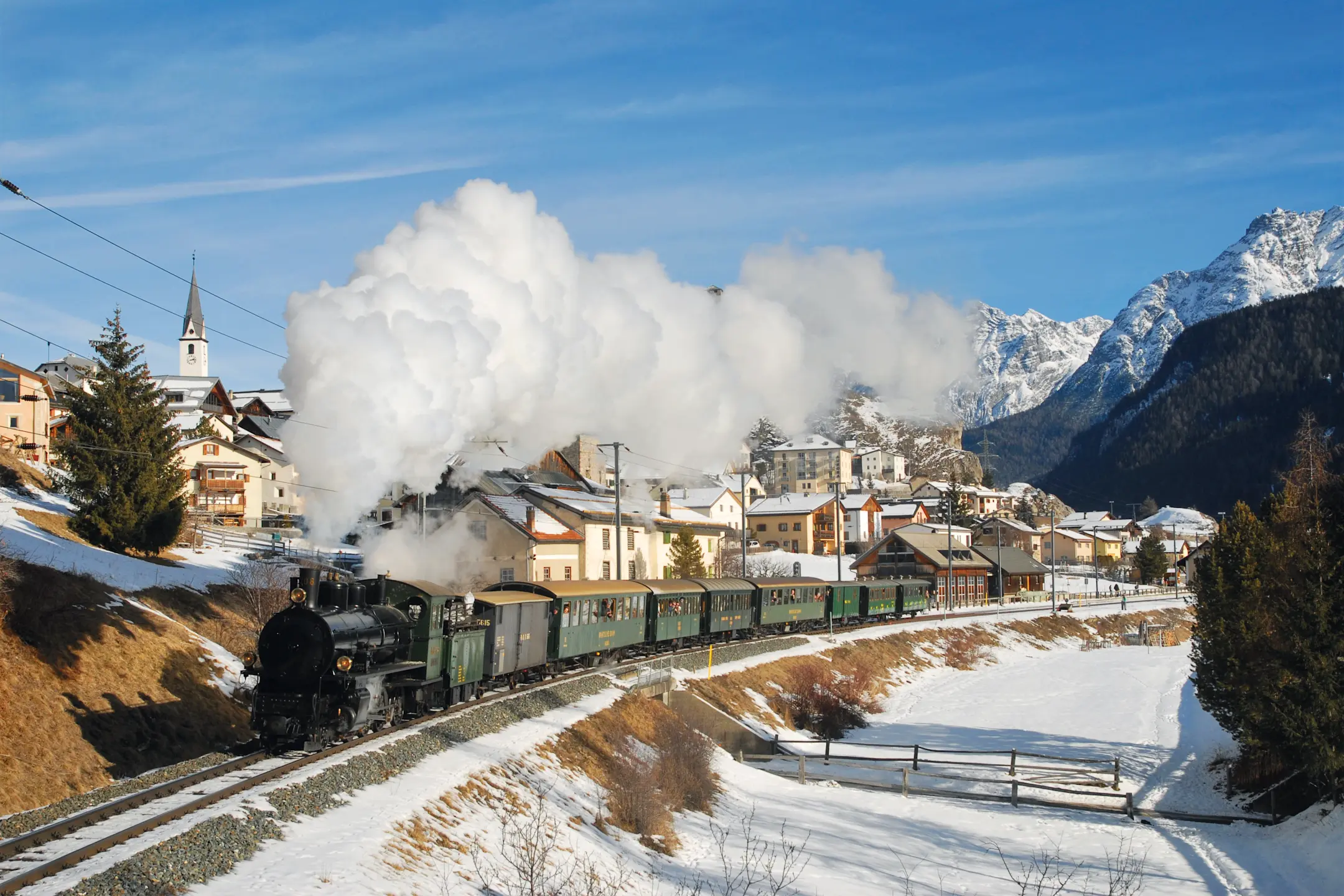 Historischer Dampfzug der RhB fährt durch das winterliche Engadiner Dorf Ardez, im Hintergrund die Gipfel des Unterengadins.