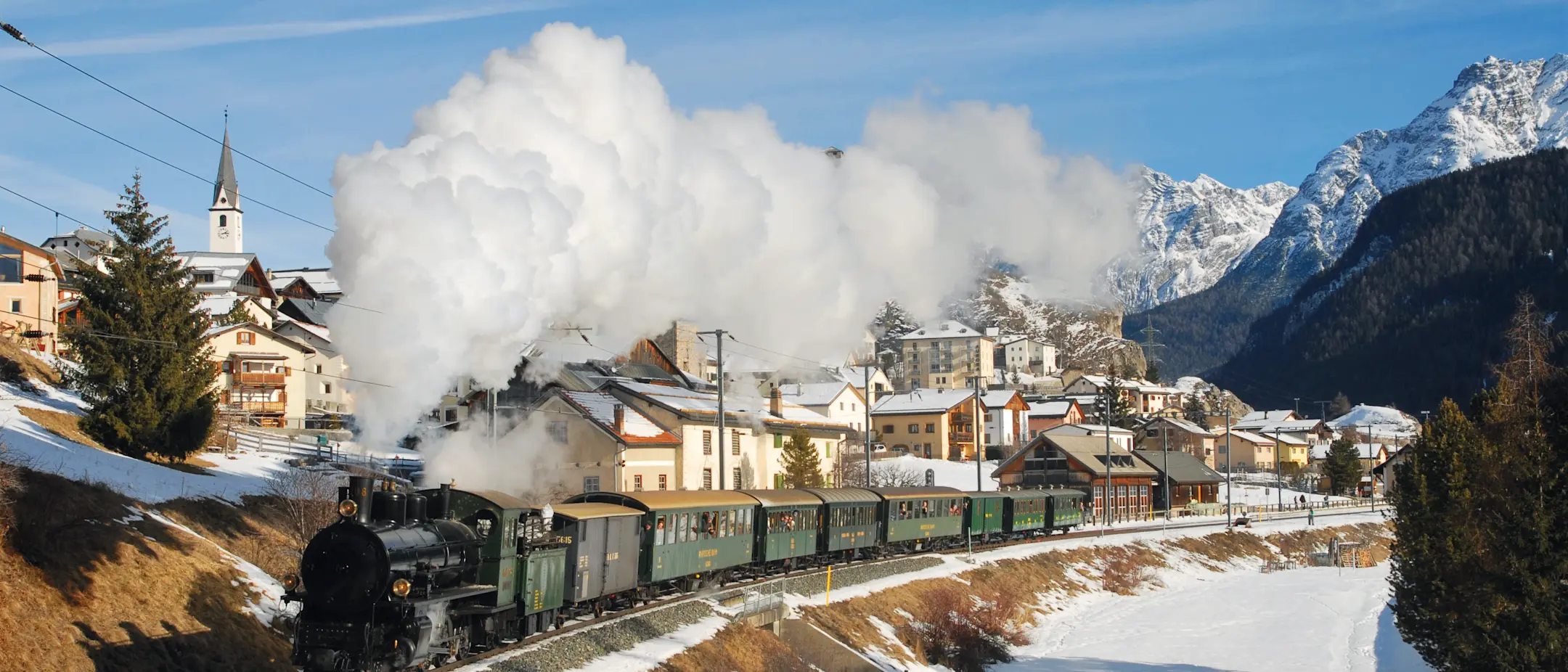 Historischer Dampfzug der RhB fährt durch das winterliche Engadiner Dorf Ardez, im Hintergrund die Gipfel des Unterengadins.