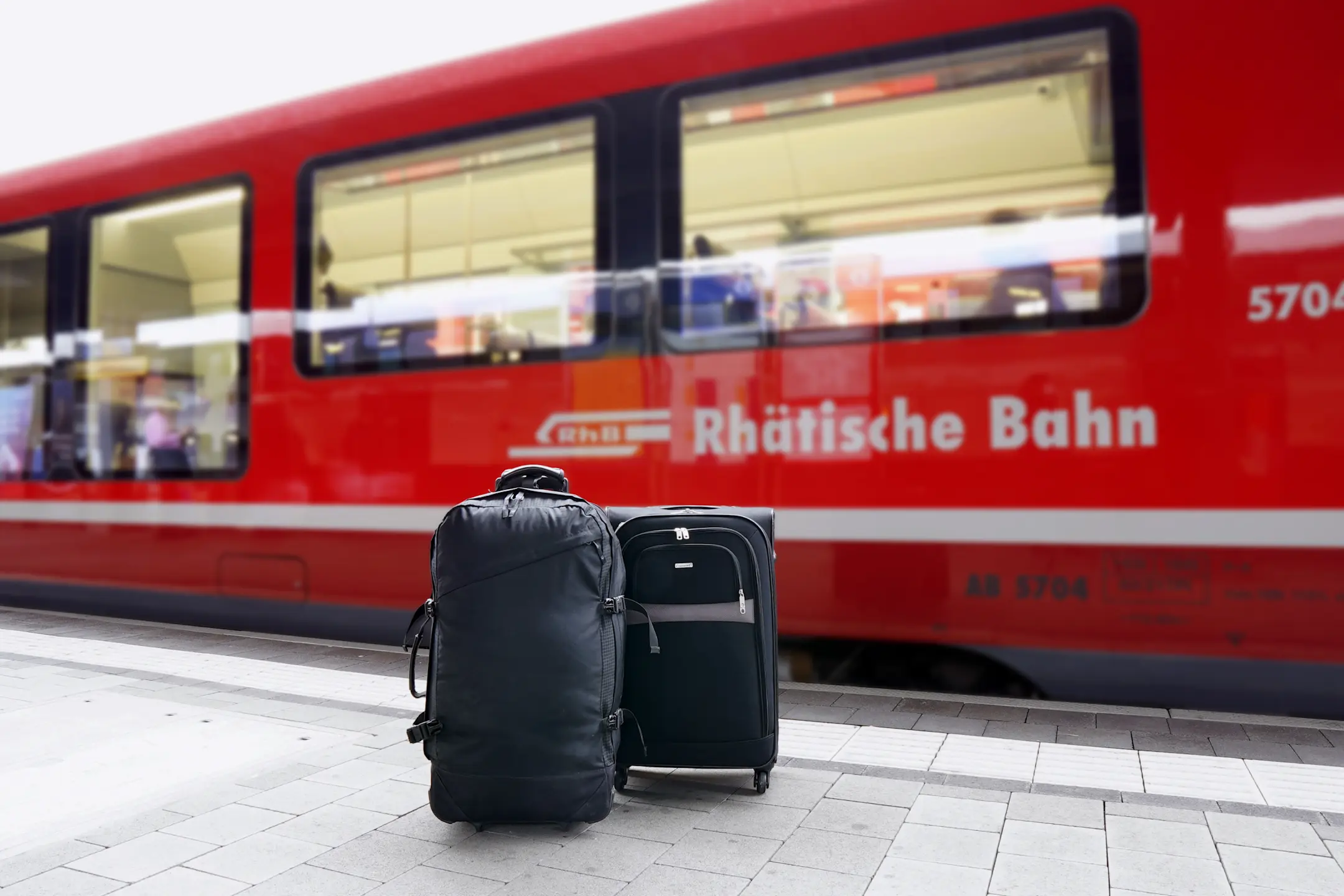 Zwei Koffer stehen auf einem Bahnhofsbahnsteig vor einem roten Zug der Rhätischen Bahn, bereit für den Gepäckservice.