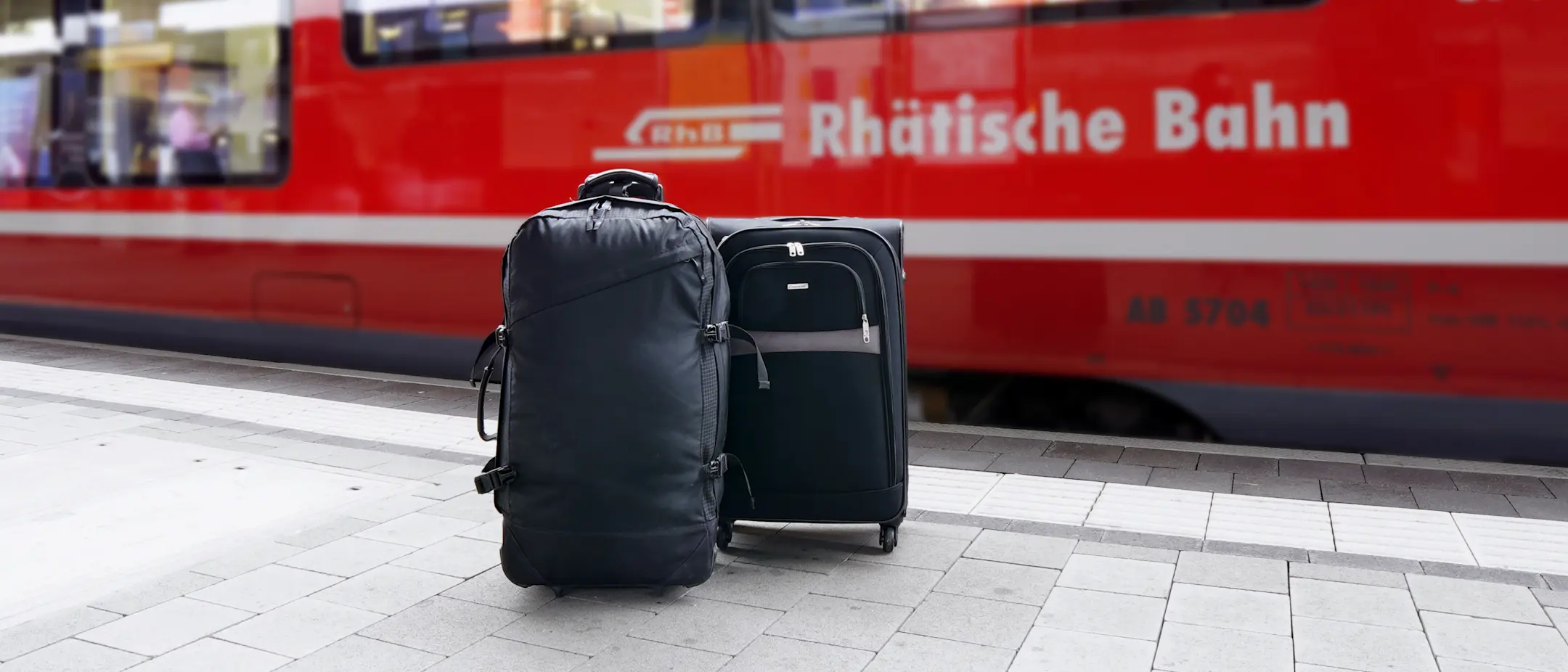 Zwei Koffer stehen auf einem Bahnhofsbahnsteig vor einem roten Zug der Rhätischen Bahn, bereit für den Gepäckservice.