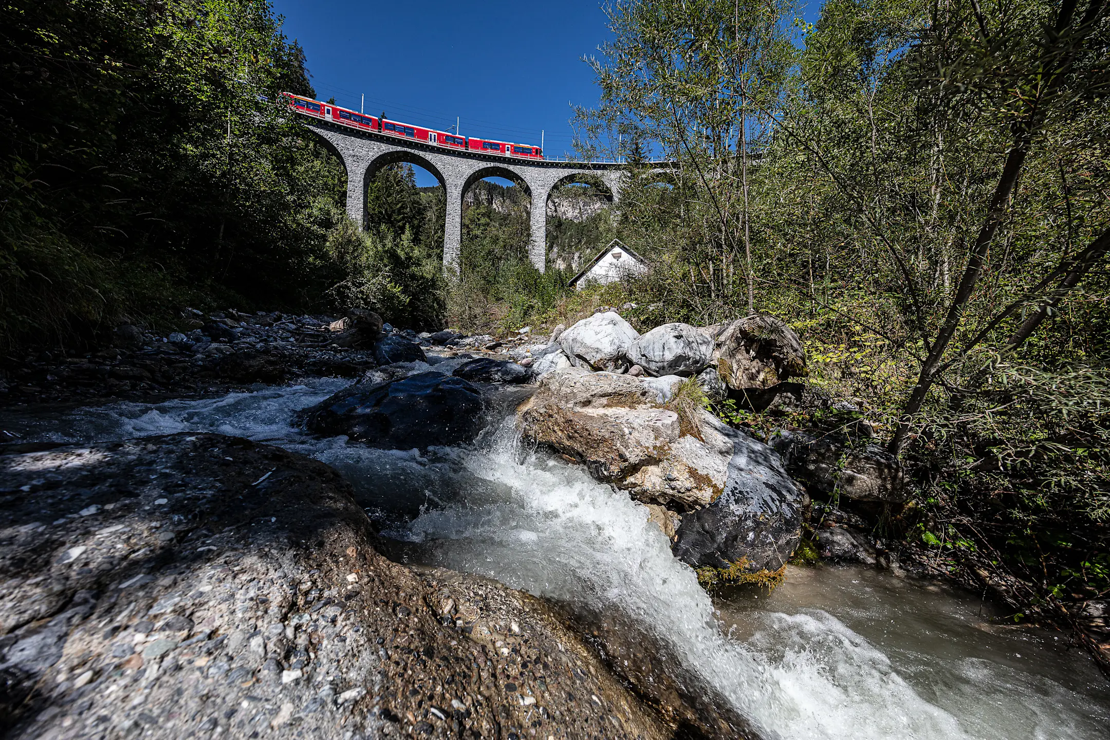 Allegra-Triebzug auf dem Schmittnertobelviadukt im Sommer mit Bach im Vordergrund.