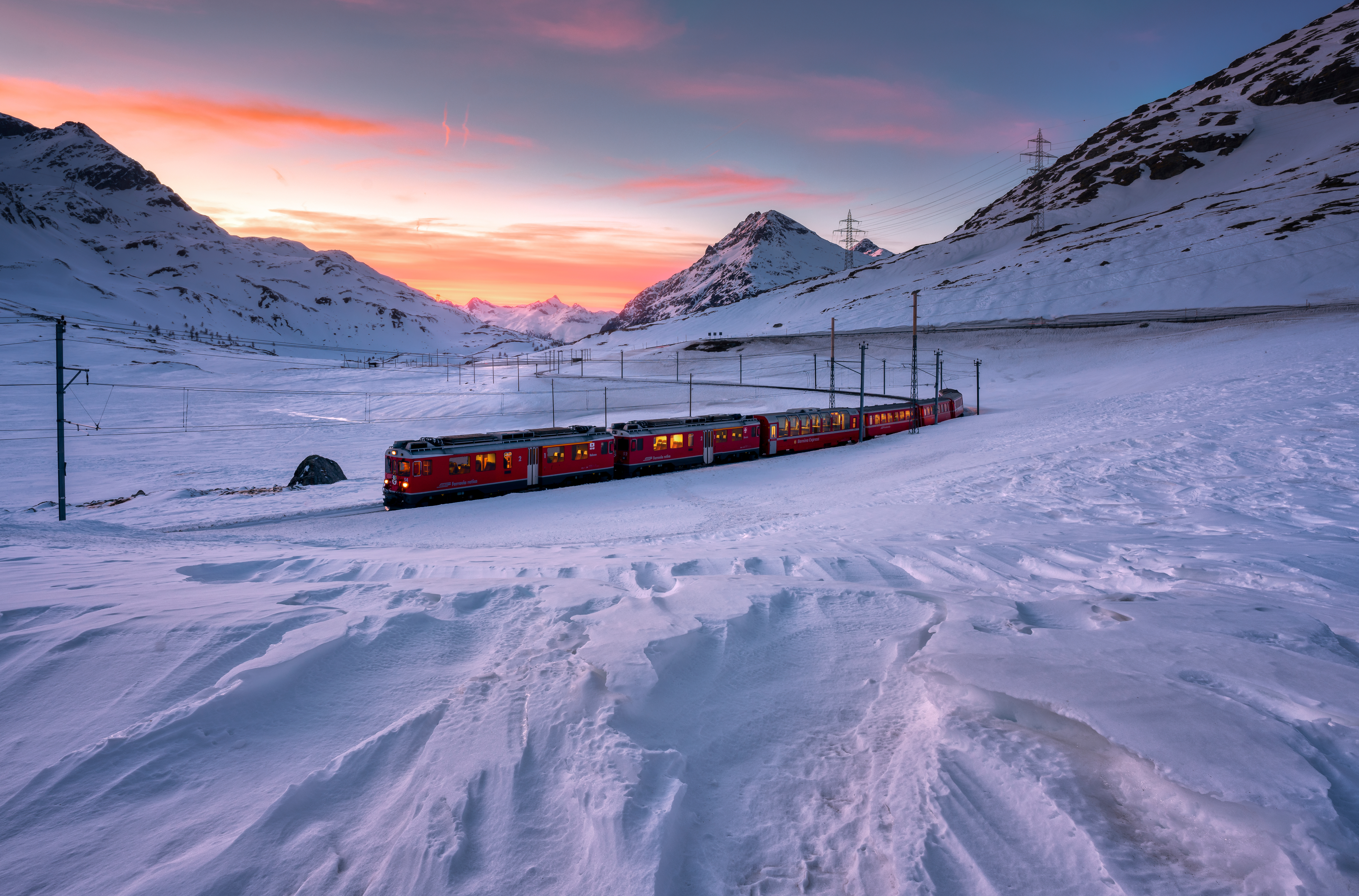 A RhB regional train travels over the snow-covered Bernina Pass in winter, with mountains and a sunset in the background.