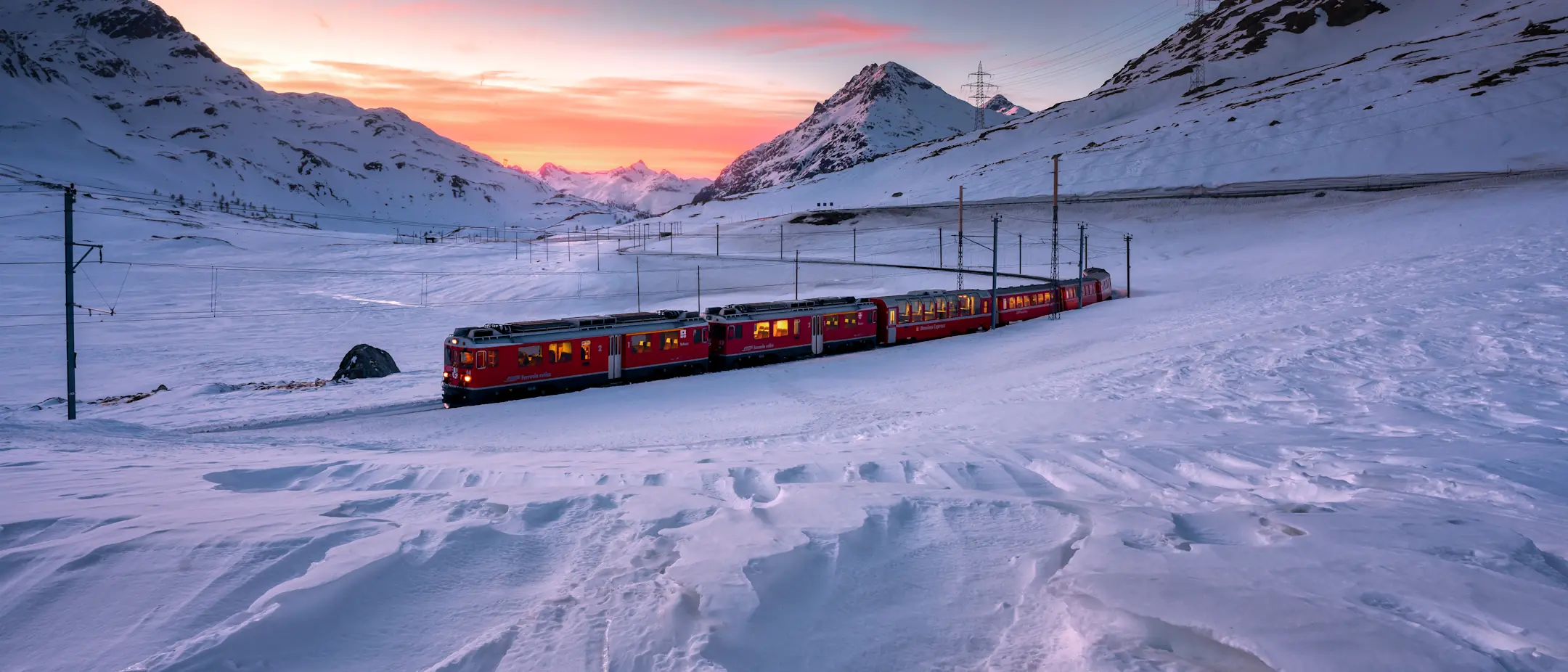 Ein Regionalzug der RhB fährt im Winter über den schneebedeckten Berninapass, im Hintergrund Berge und ein Sonnenuntergang