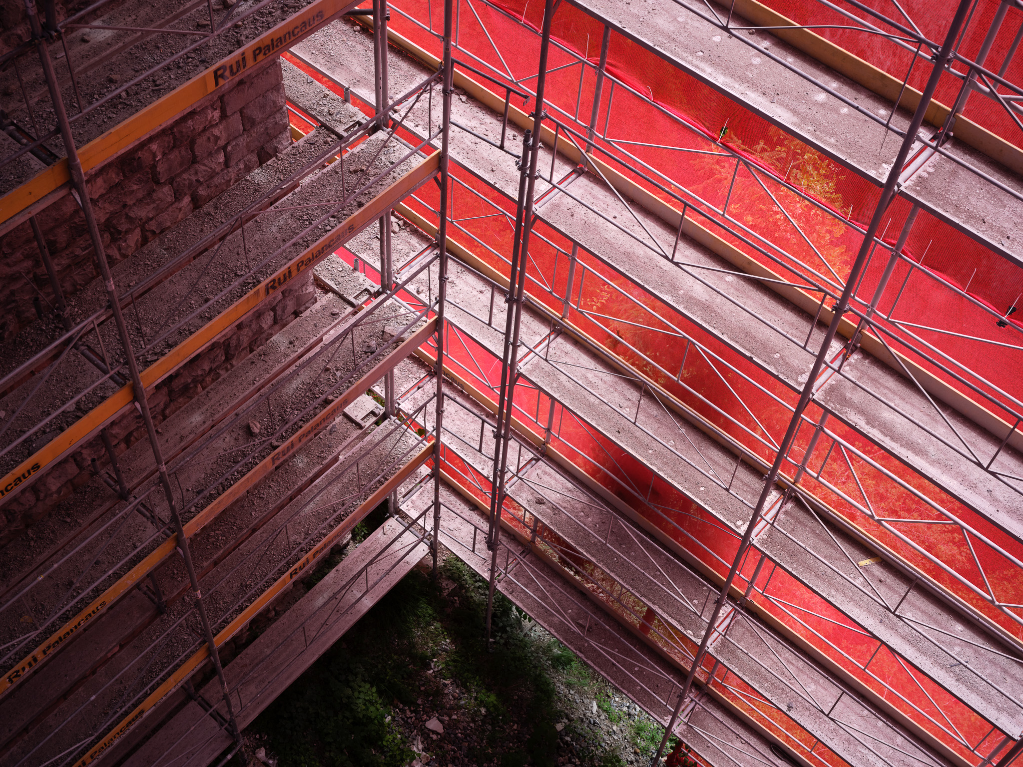 Mehrstöckiges Gerüst mit roter Schutzverkleidung an einer Steinstruktur während Bau‑ bzw. Sanierungsarbeiten.