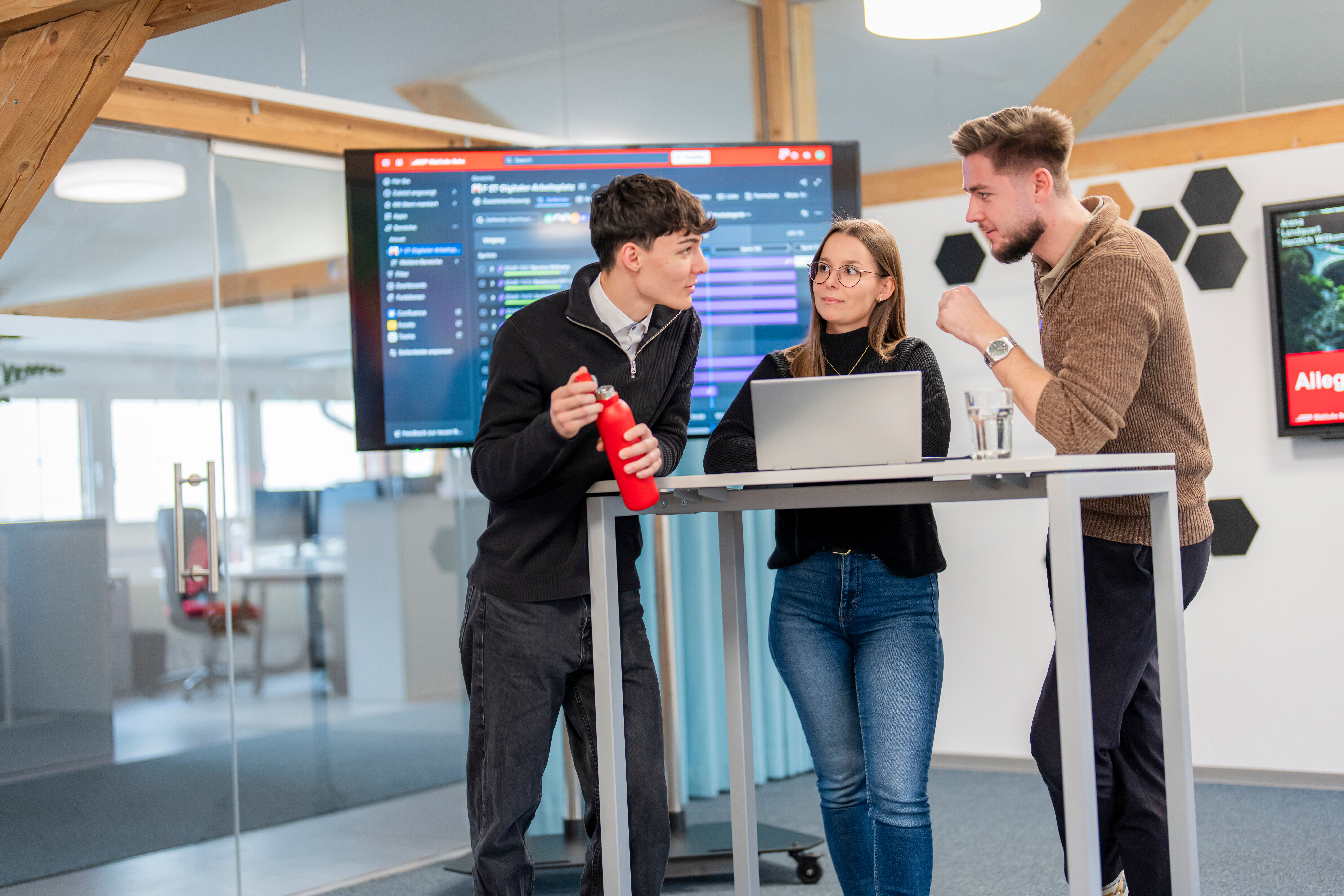 Drei IT Mitarbeitende bei einer Besprechung mit Notebook und Bildschirm im Hintergrund