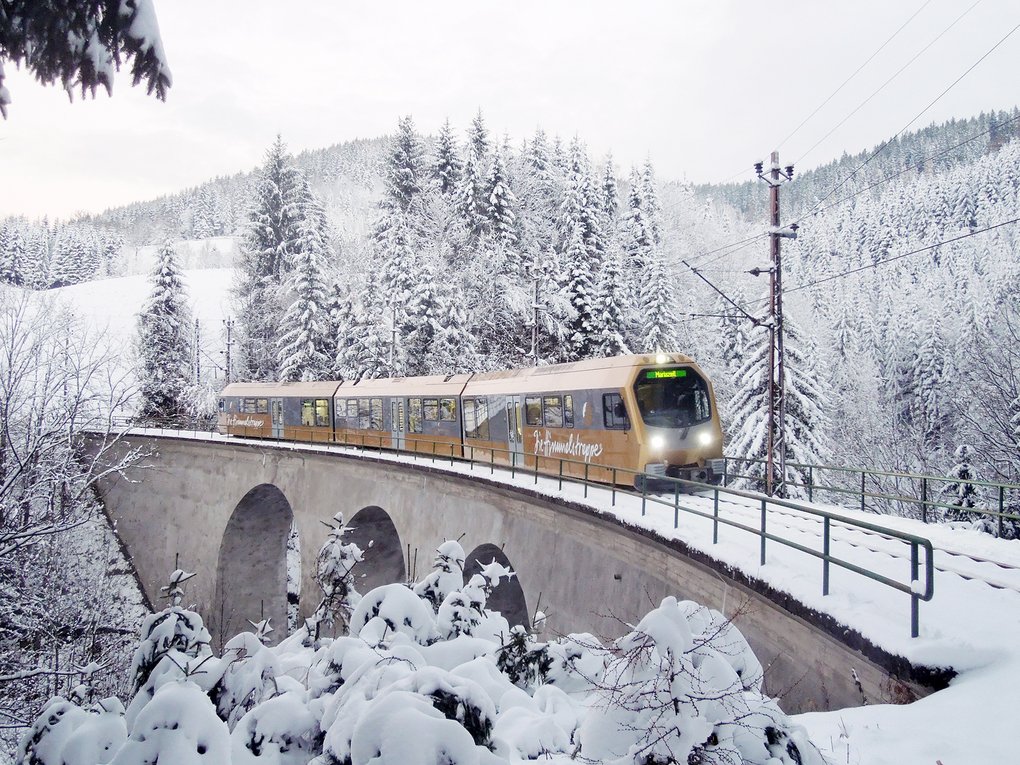 A golden train of the Mariazellerbahn railway travels over a viaduct through a snow-covered forest landscape in Austria.