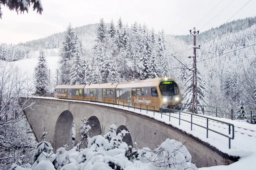 Ein goldfarbener Zug der Mariazellerbahn fährt über ein Viadukt durch eine tief verschneite Waldlandschaft in Österreich.