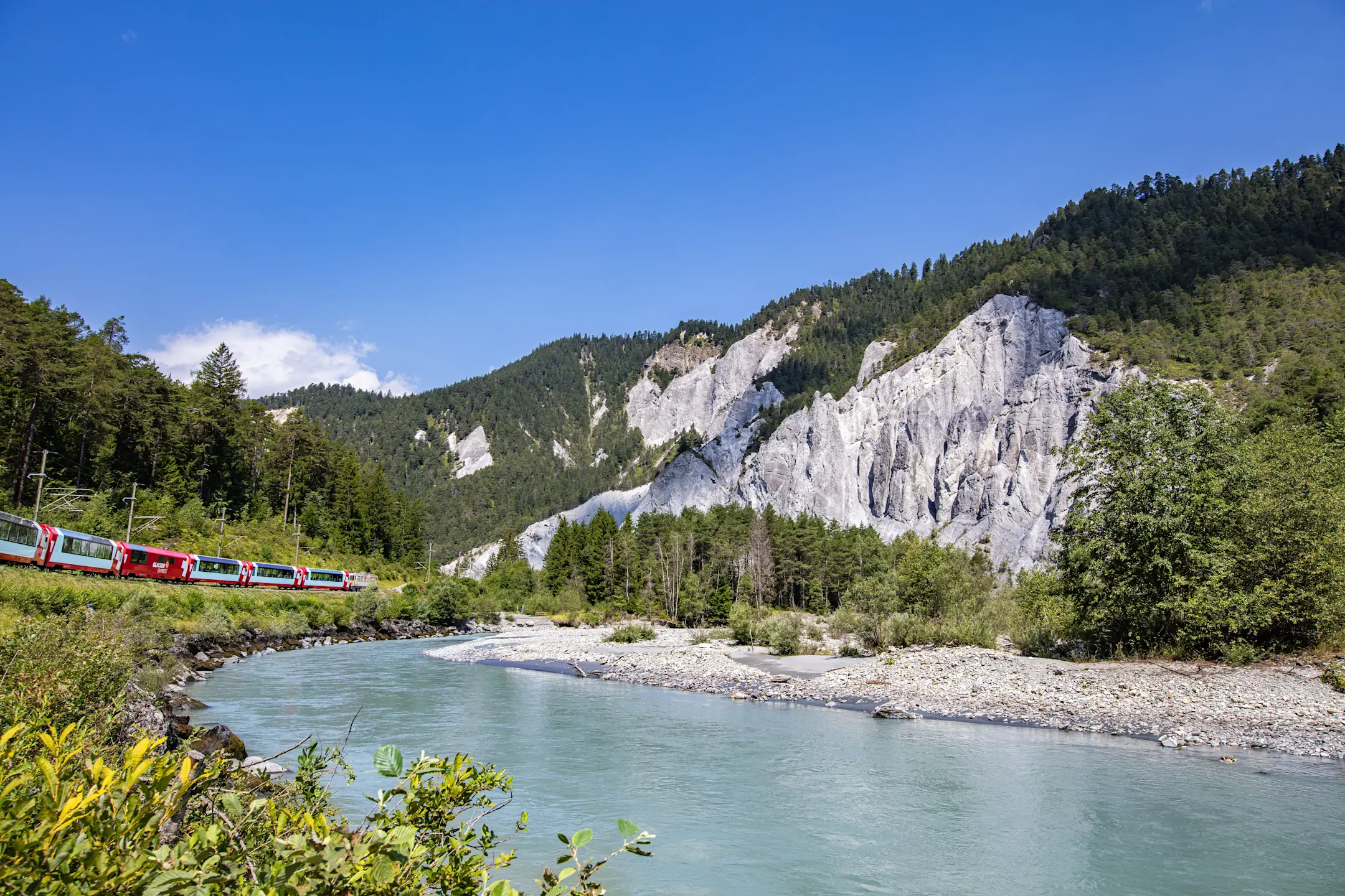 Ein Zug schlängelt sich durch die Rheinschlucht entlang des türkisfarbenen Rheins, eingerahmt von Felsen und Wäldern.