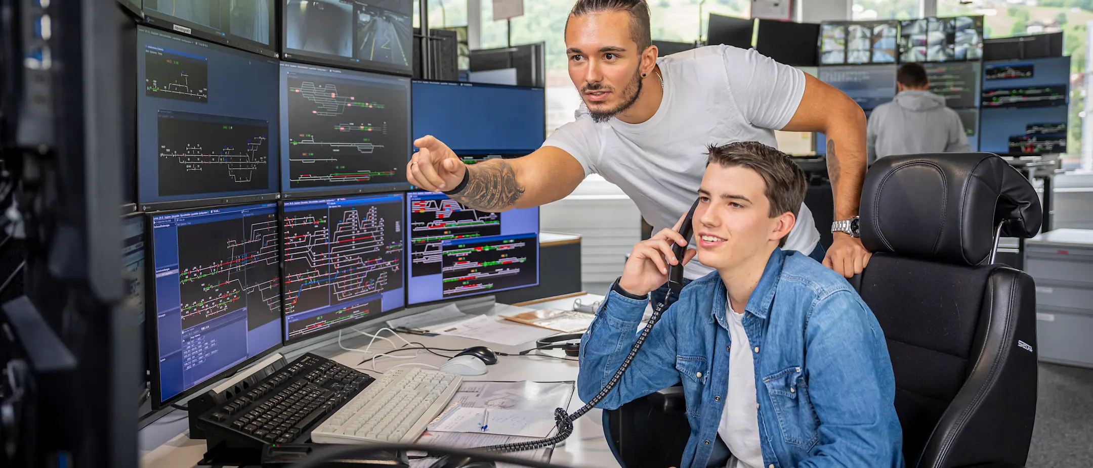 Ein Zugverkehrsleiter zeigt einem jungen Kollegen in der Betriebszentrale ein Detail auf einem Monitor.