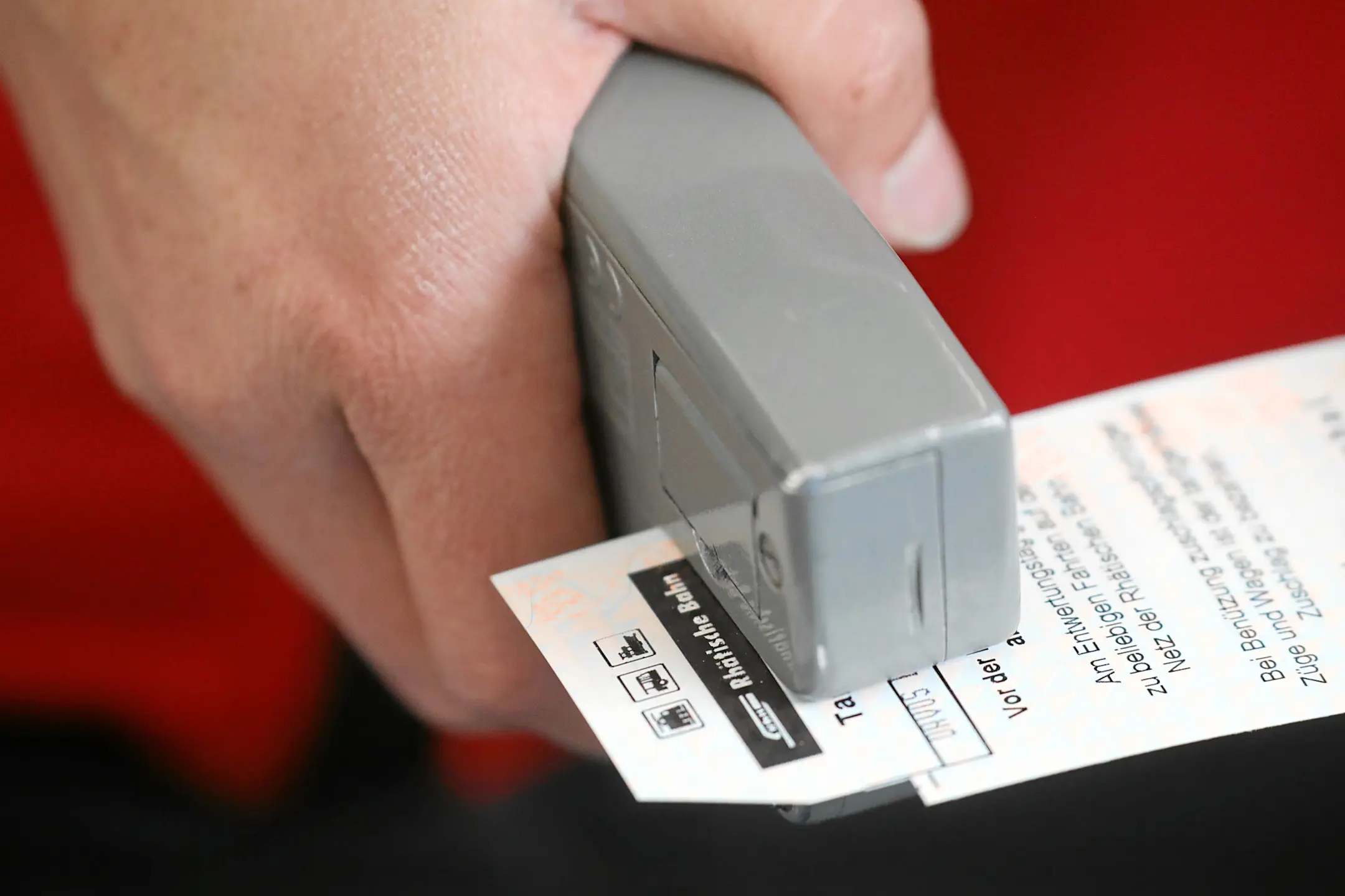 Nahaufnahme einer Hand, die ein Rhätische-Bahn-Ticket mit einem Entwerter stempelt.