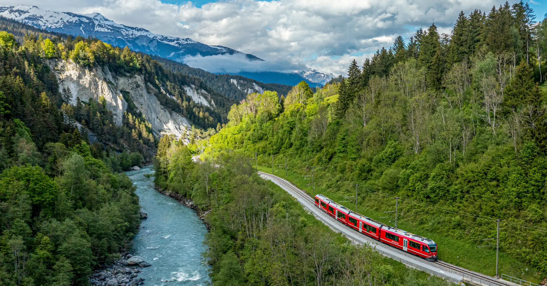 Ein roter Zug fährt entlang des Rheins durch die tief eingeschnittene, bewaldete Rheinschlucht mit Blick Alpen.