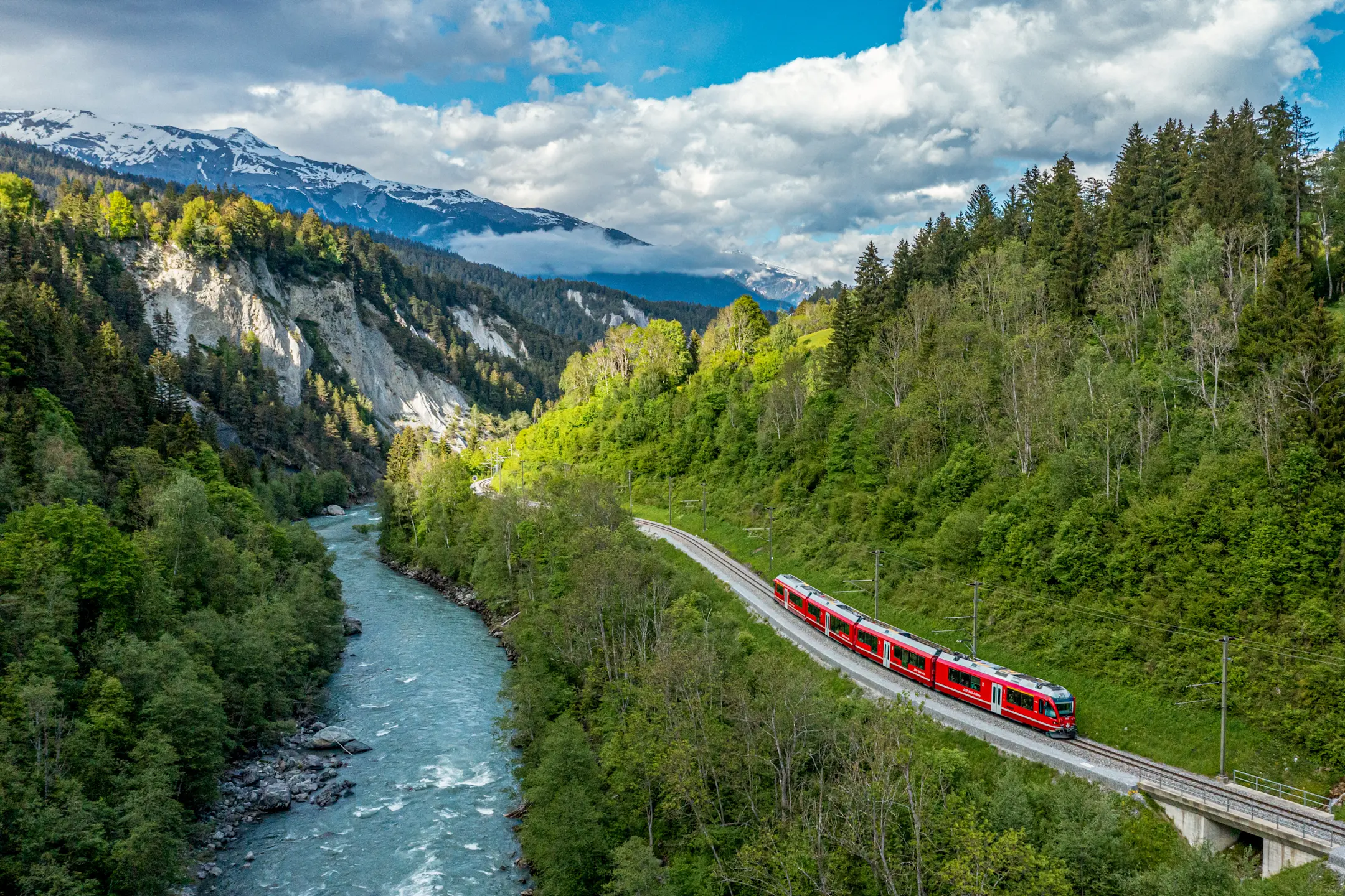 Ein roter Zug fährt entlang des Rheins durch die tief eingeschnittene, bewaldete Rheinschlucht mit Blick Alpen.
