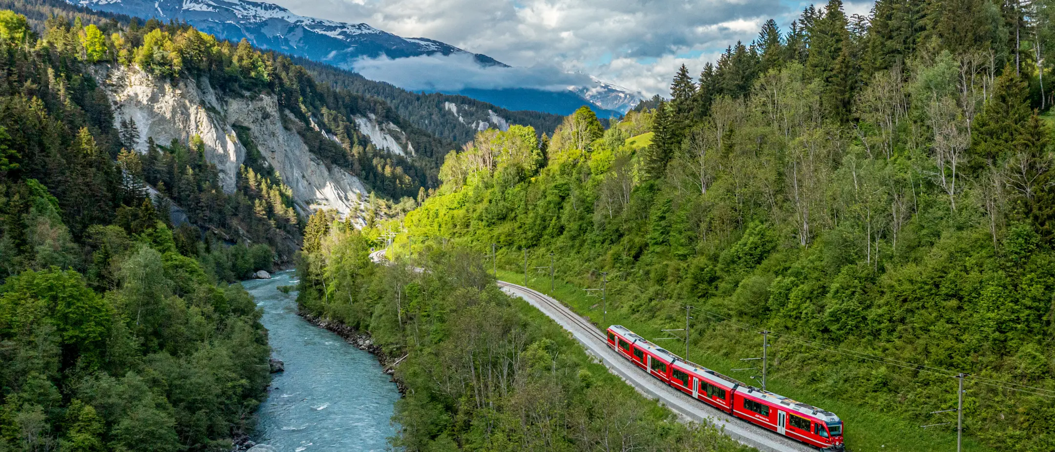 Ein roter Zug fährt entlang des Rheins durch die tief eingeschnittene, bewaldete Rheinschlucht mit Blick Alpen.