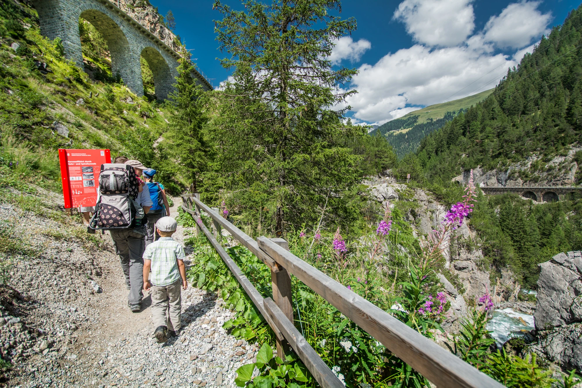 Eine Familie wandert auf dem Bahnerlebnisweg Albula entlang eines Viadukts der Rhätischen Bahn inmitten einer Berglandschaft.