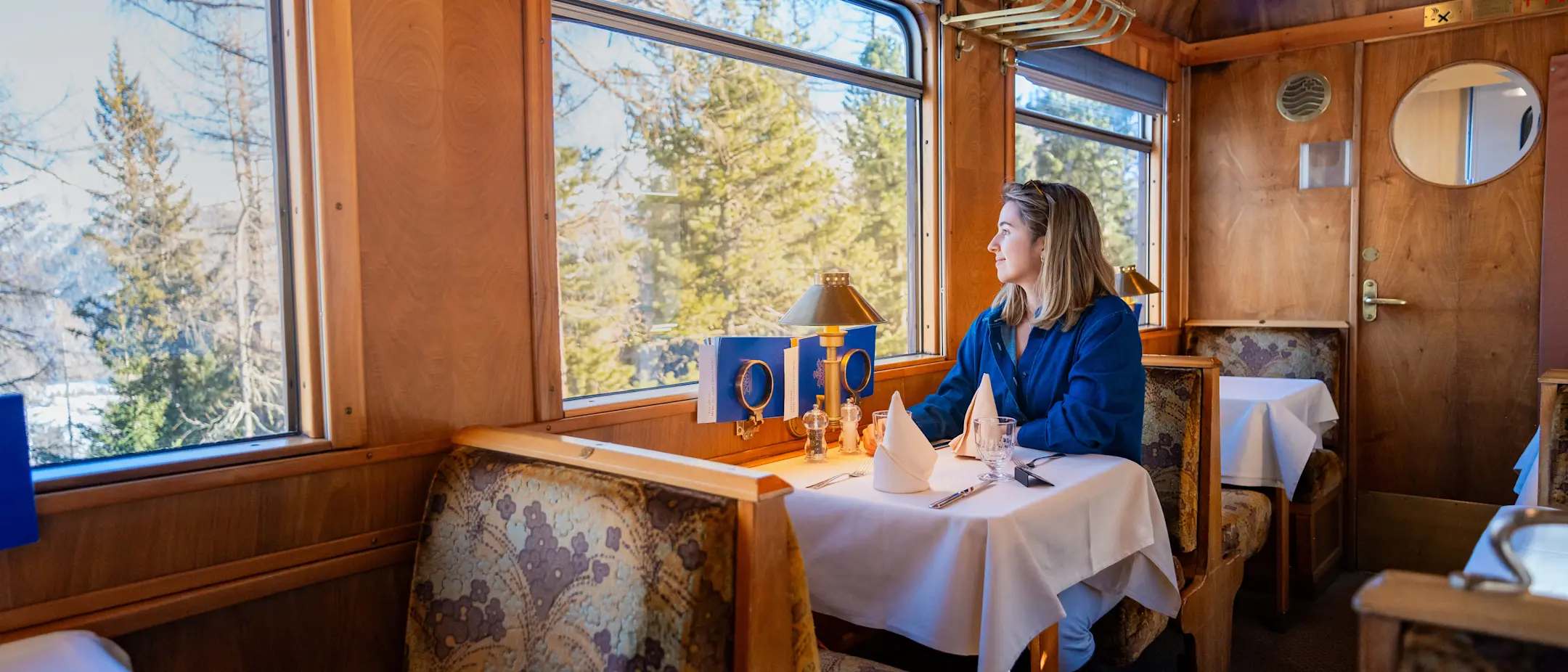 Frau sitzt an einem weiß gedeckten Tisch im historischen Speisewagen Gourmino und genießt die Aussicht durch das Zugfenster.
