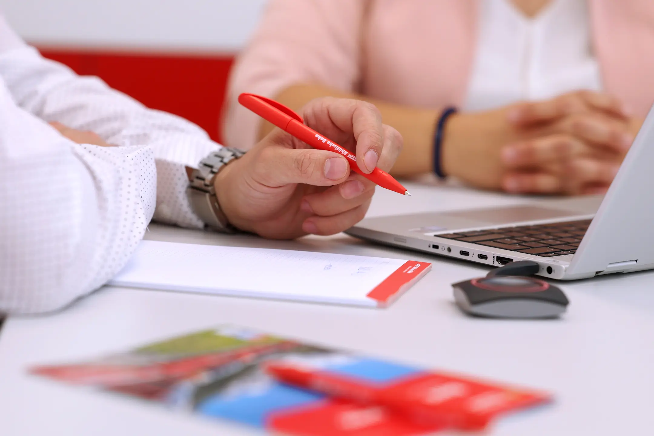 Zwei Personen bei einer Besprechung, im Fokus ein roter Kugelschreiber mit RhB-Logo, Notizblock und Laptop auf dem Tisch.