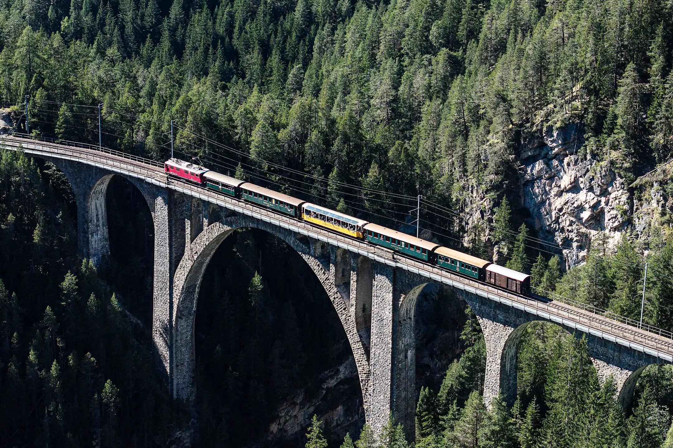 Ein historischer Zug mit nostalgischen Waggons überquert ein hohes Viadukt mitten durch einen Bergwald in Graubünden.