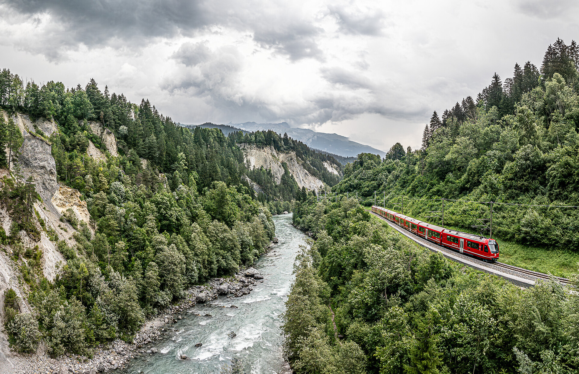 Ein roter Zug der Rhätischen Bahn fährt entlang der Rheinschlucht, flankiert vom Fluss und hellen Felswänden.