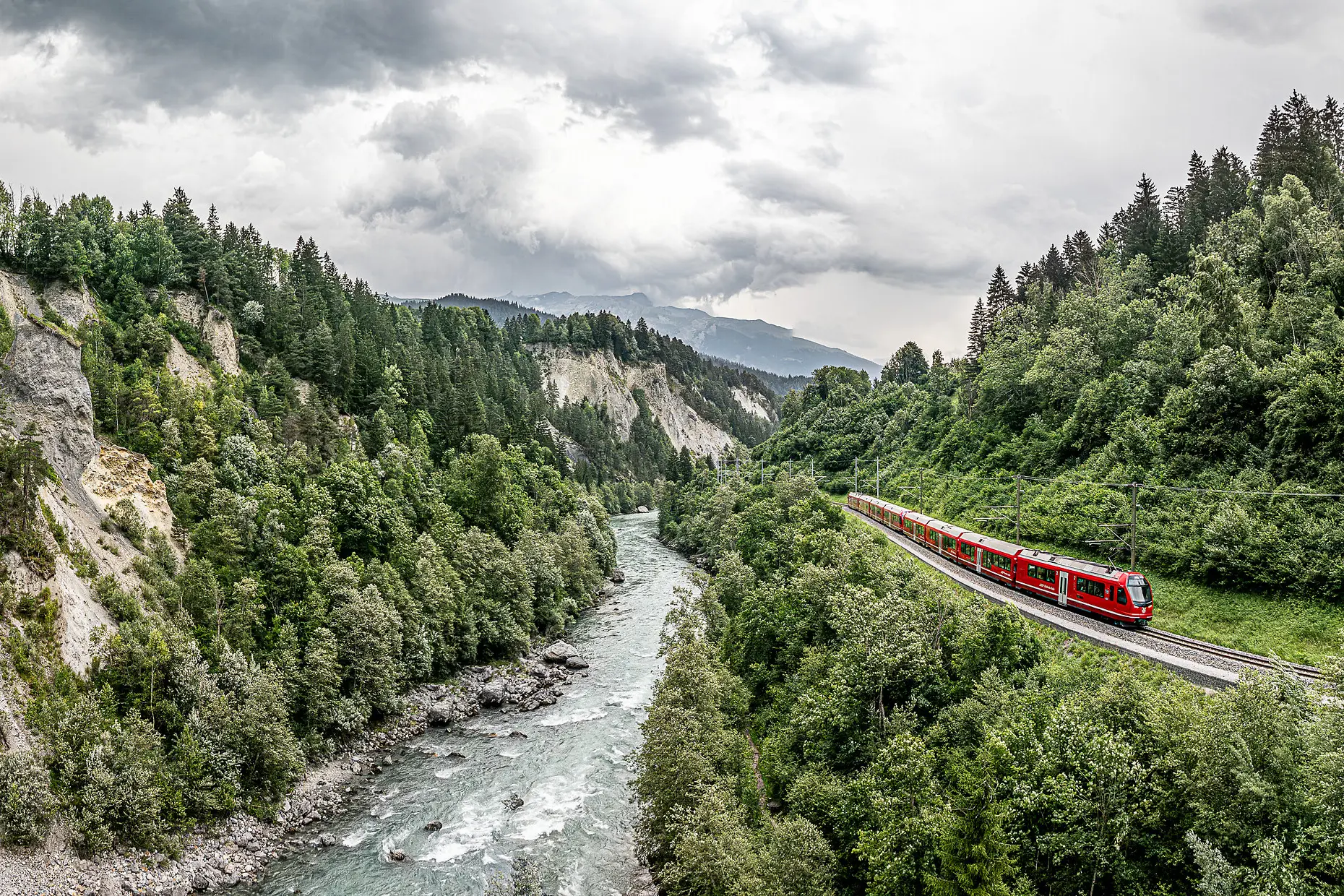 Ein roter Zug der Rhätischen Bahn fährt entlang der Rheinschlucht, flankiert vom Fluss und hellen Felswänden.