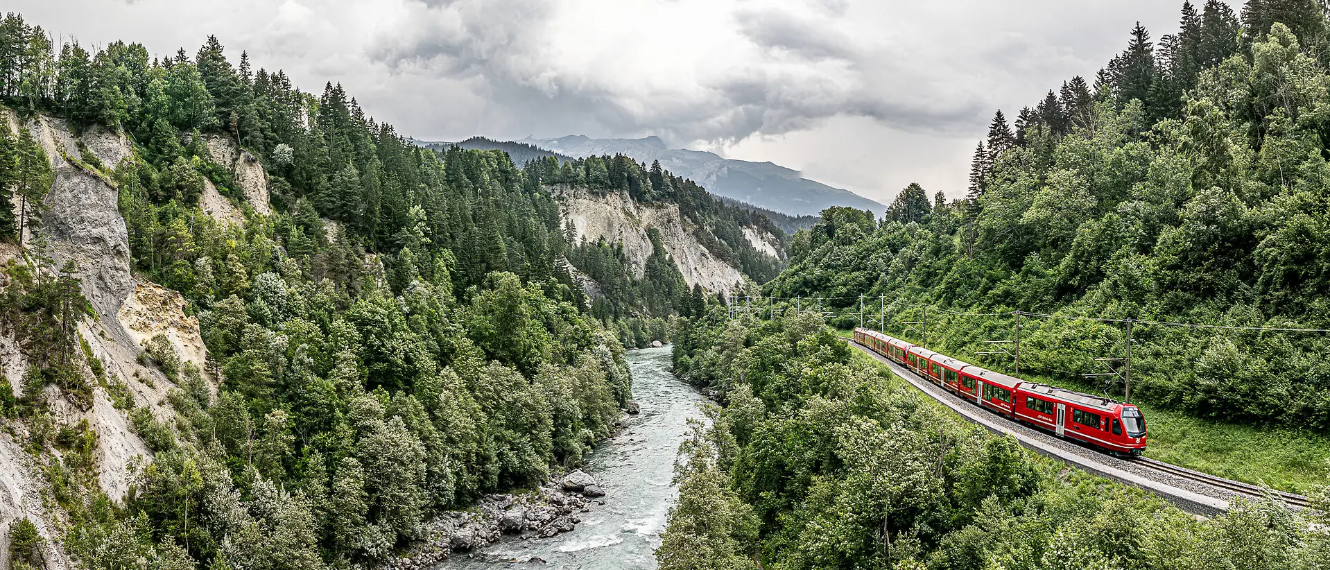 Ein roter Zug der Rhätischen Bahn fährt entlang der Rheinschlucht, flankiert vom Fluss und hellen Felswänden.