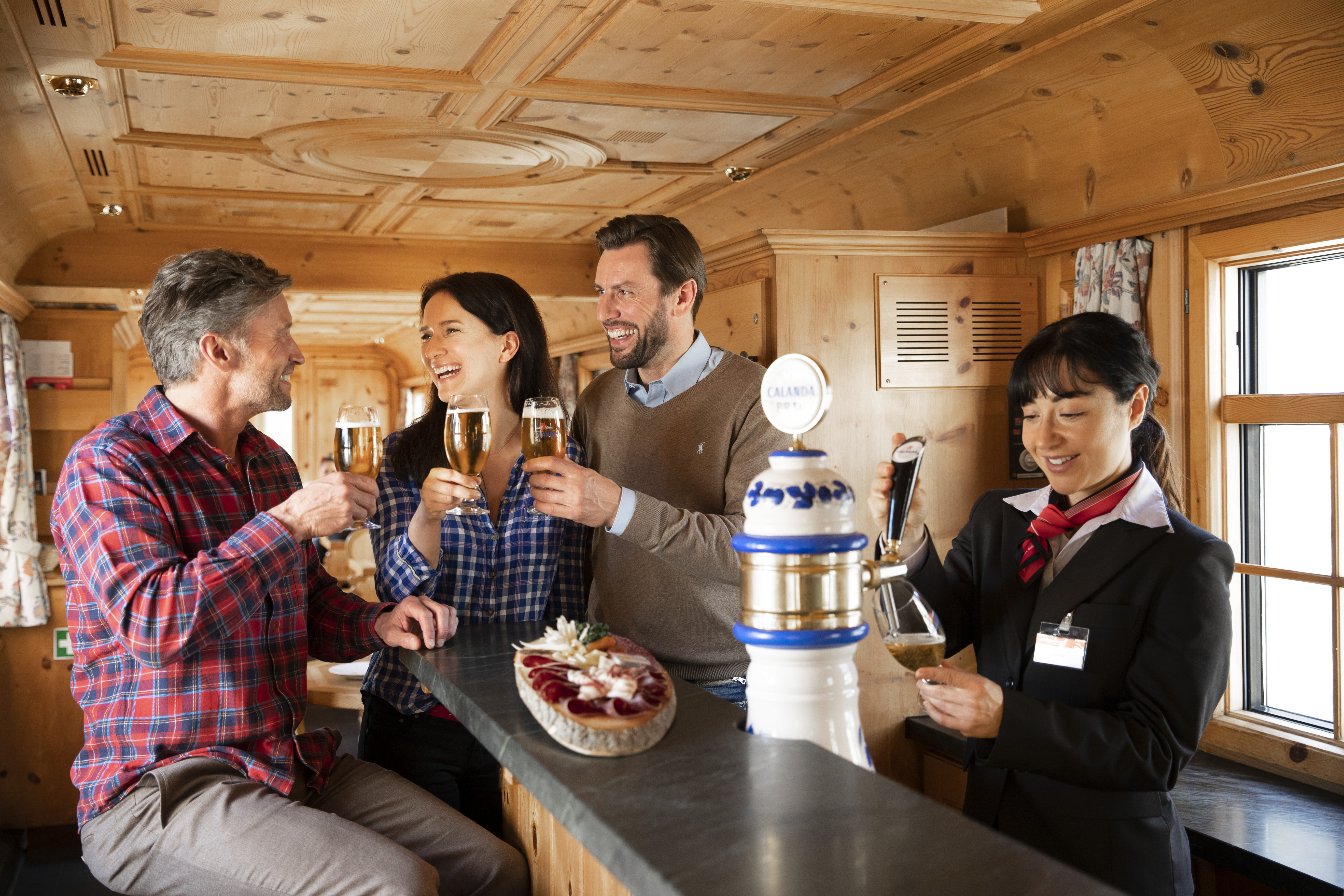 Vier fröhliche Menschen stoßen mit Biergläsern in einem holzverkleideten Barwagen der Rhätischen Bahn an.