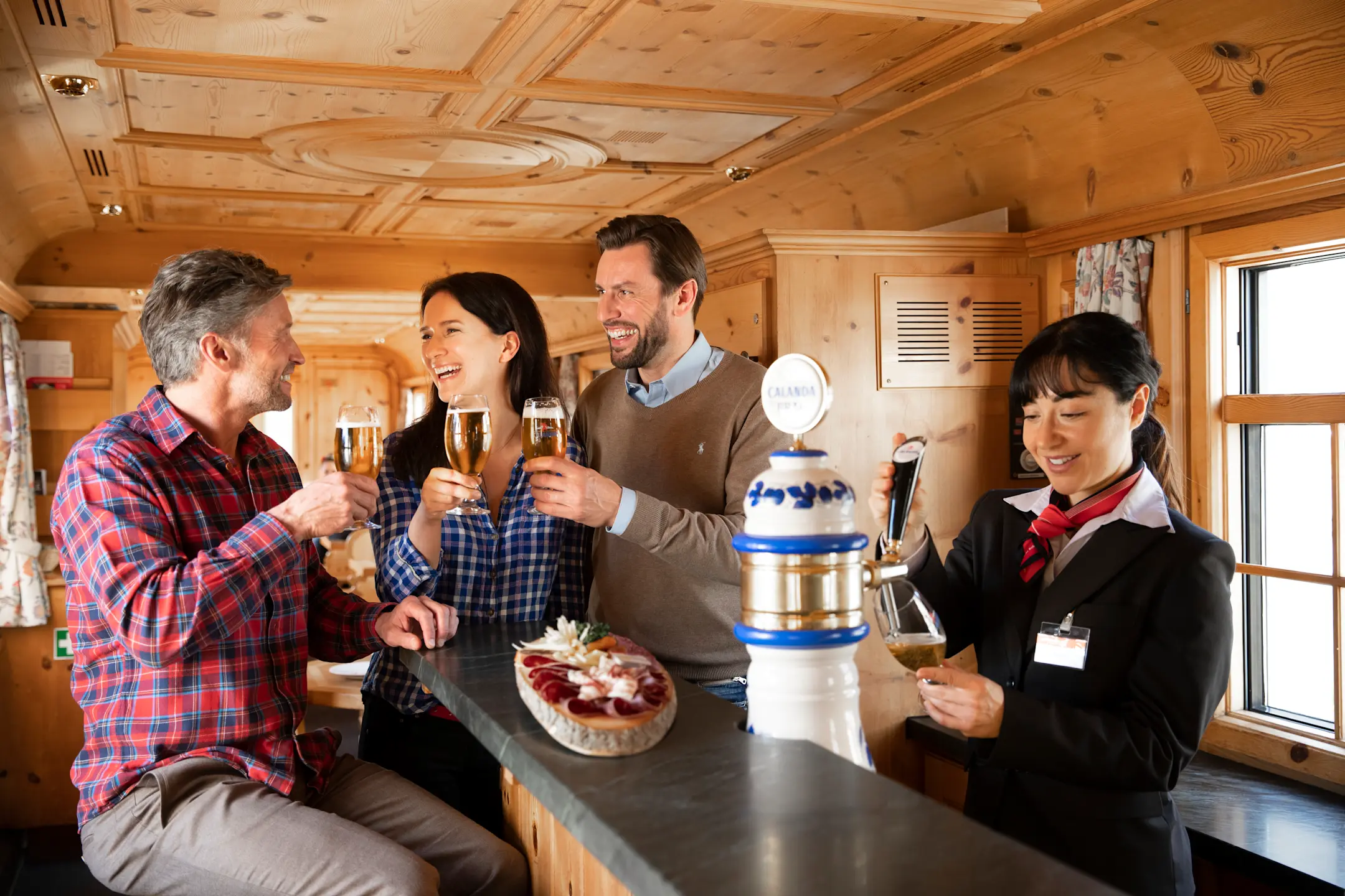 Vier fröhliche Menschen stoßen mit Biergläsern in einem holzverkleideten Barwagen der Rhätischen Bahn an.