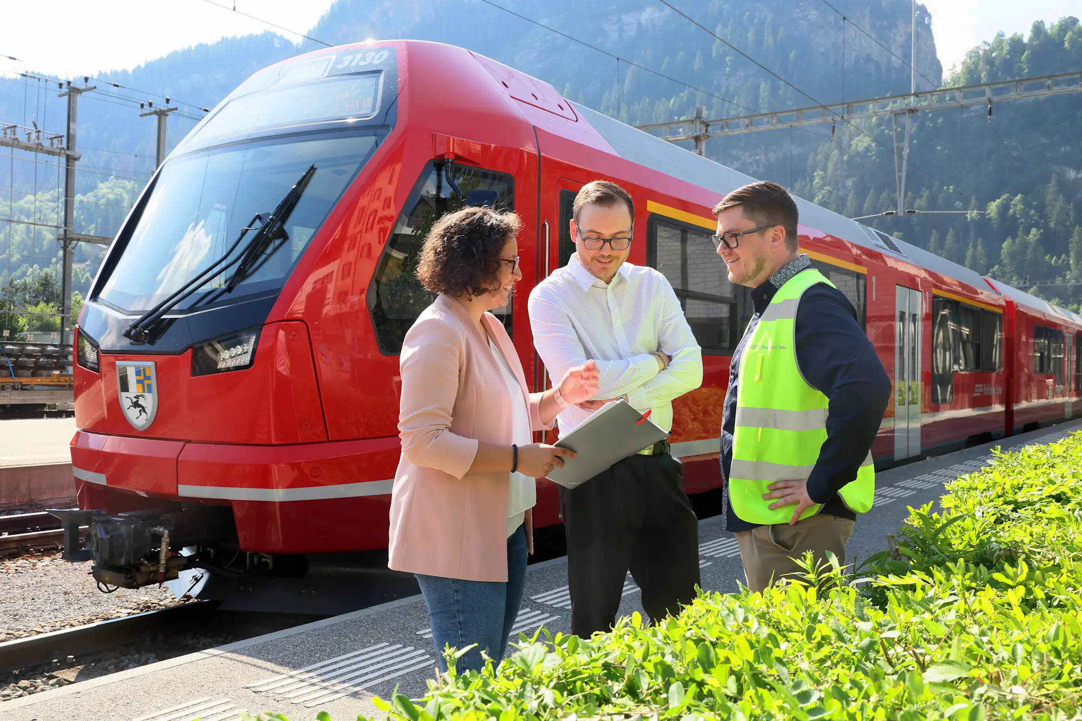 Drei RhB-Mitarbeitende besprechen sich auf dem Bahnsteig vor einem roten Capricorn-Triebzug – bei Sonne und Alpenkulisse.