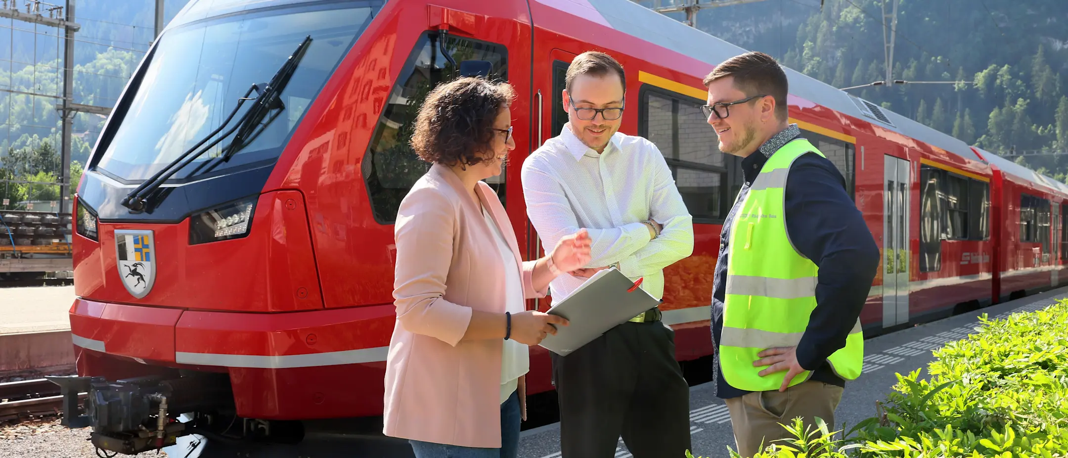 Drei RhB-Mitarbeitende besprechen sich auf dem Bahnsteig vor einem roten Capricorn-Triebzug – bei Sonne und Alpenkulisse.