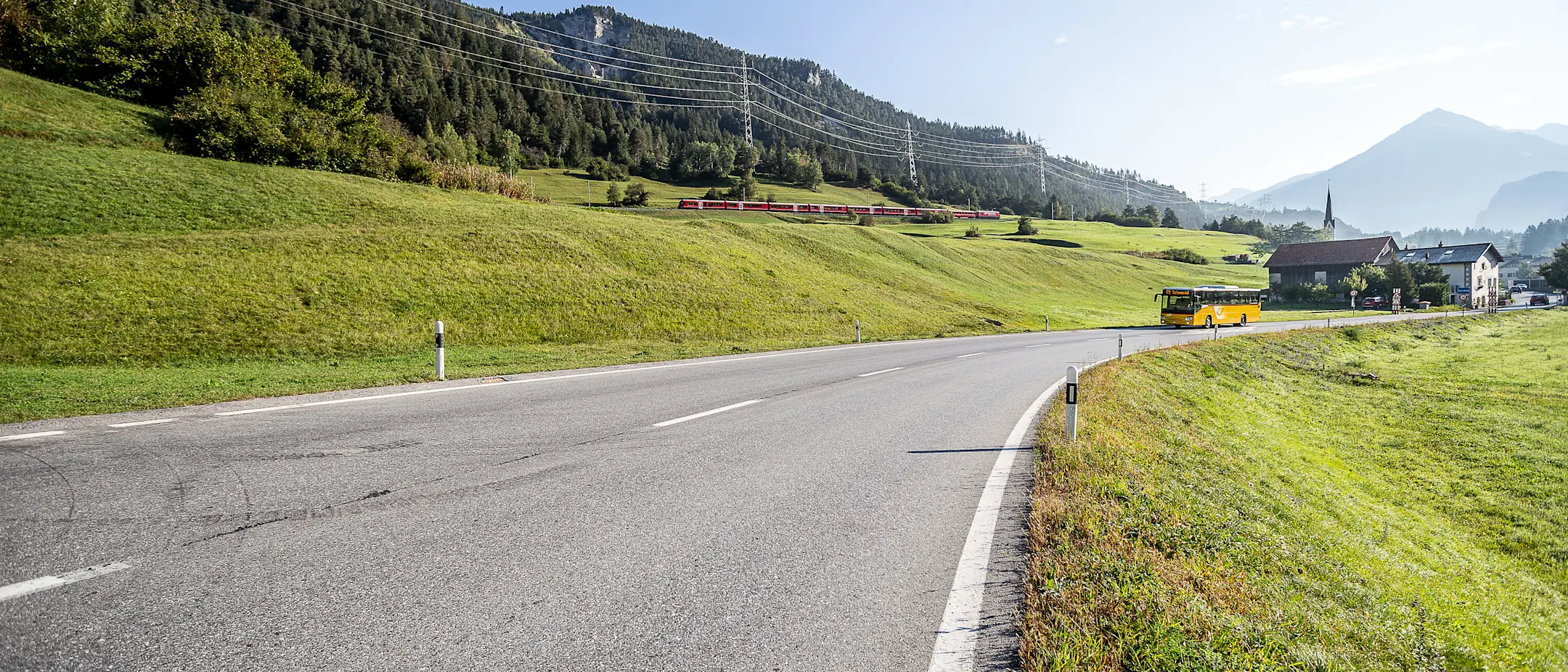Postauto auf der Strasse und Interregiozug im Hintergrund
