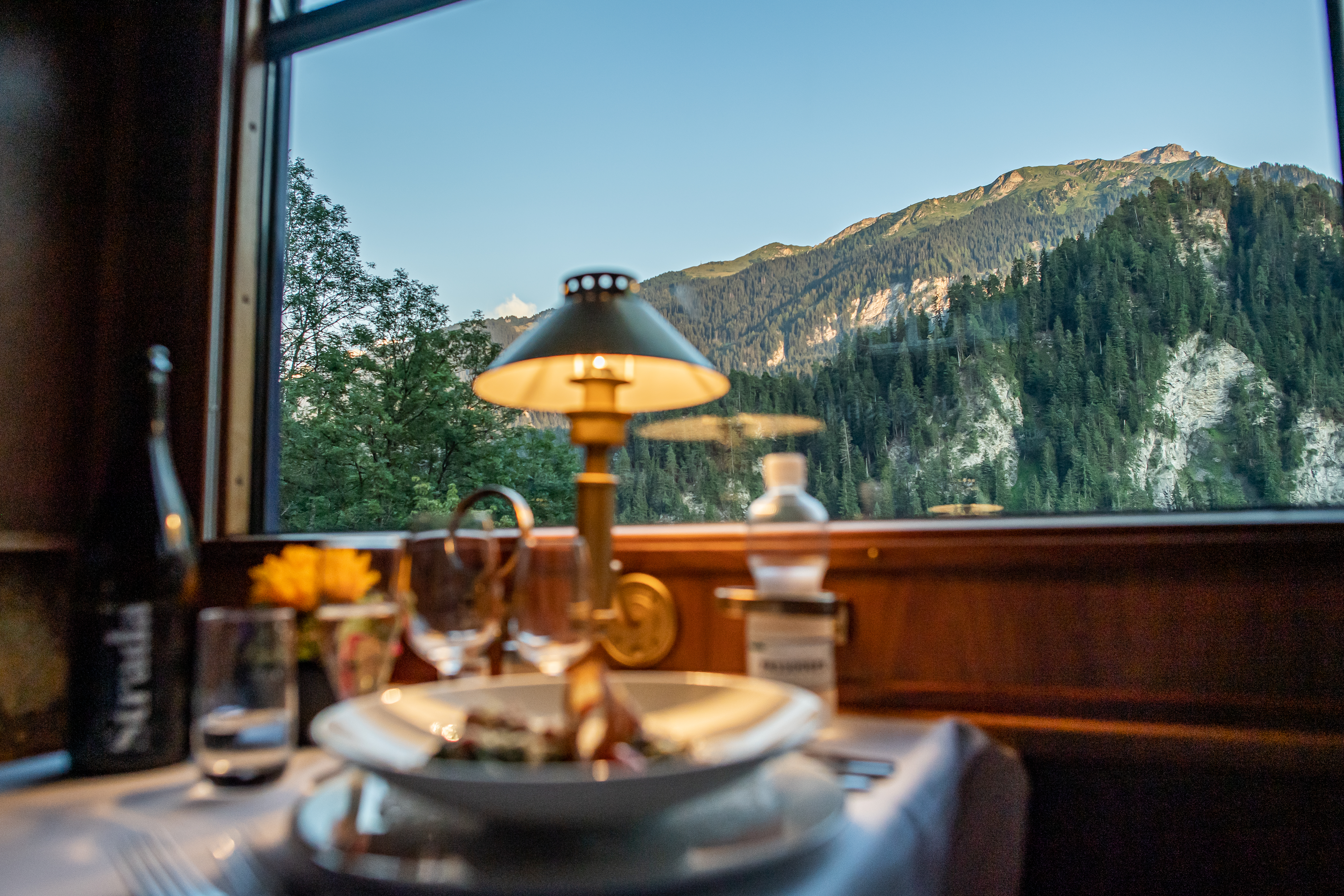 Stilvoll gedeckter Tisch mit einer warmen Lampe im Speisewagen eines Zuges. Eine malerische Alpenlandschaft im Hintergrund..