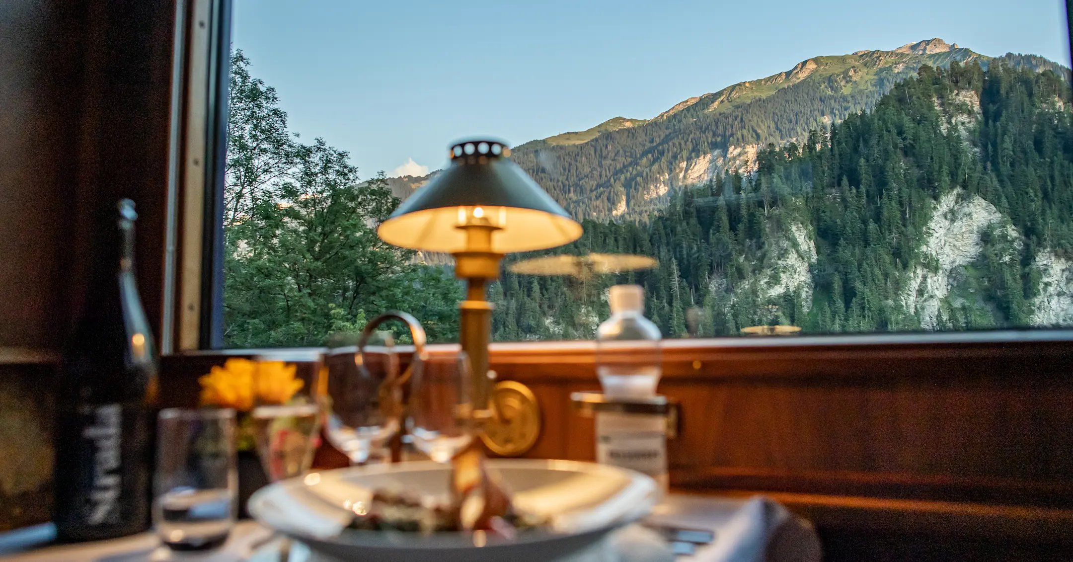 Stilvoll gedeckter Tisch mit einer warmen Lampe im Speisewagen eines Zuges. Eine malerische Alpenlandschaft im Hintergrund..