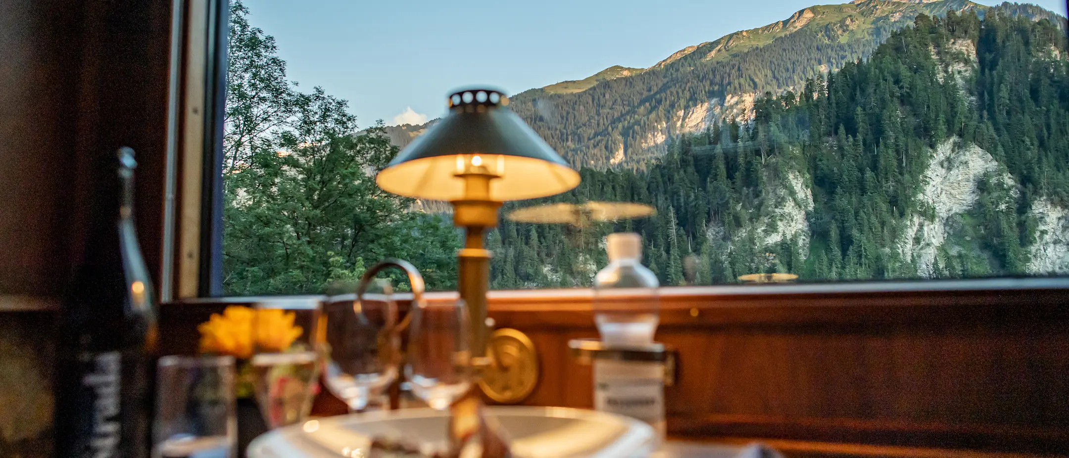 Stilvoll gedeckter Tisch mit einer warmen Lampe im Speisewagen eines Zuges. Eine malerische Alpenlandschaft im Hintergrund..