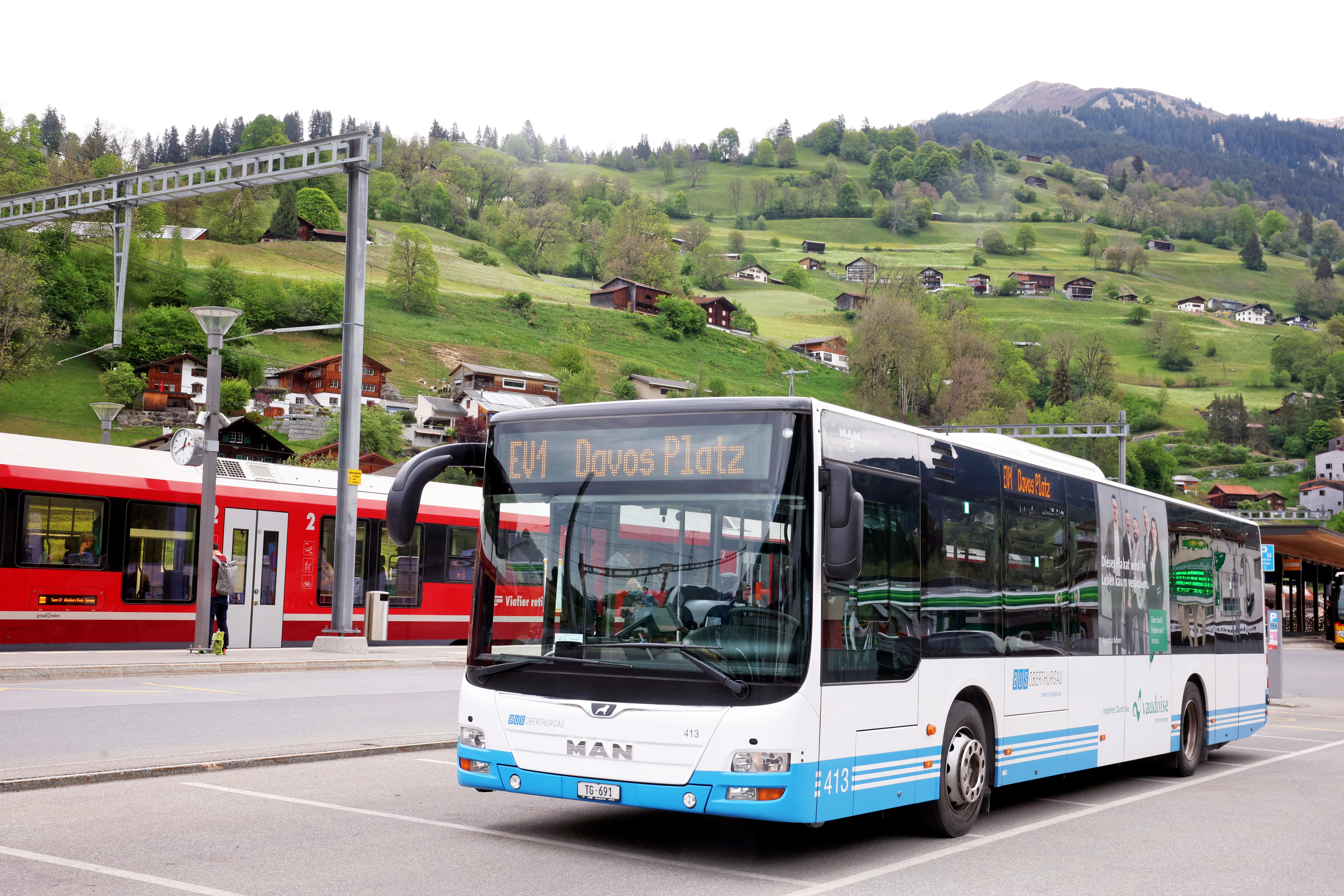 Ein hellblauer Bahnersatzbus mit dem Fahrtziel Davos Platz. Er steht in Küblis und im Hintergrund ist eine RhB-Bahn zu sehen.