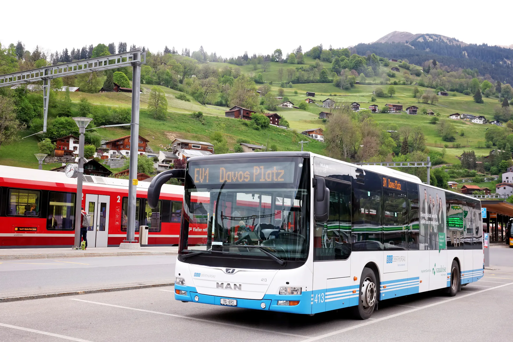 Ein hellblauer Bahnersatzbus mit dem Fahrtziel Davos Platz. Er steht in Küblis und im Hintergrund ist eine RhB-Bahn zu sehen.