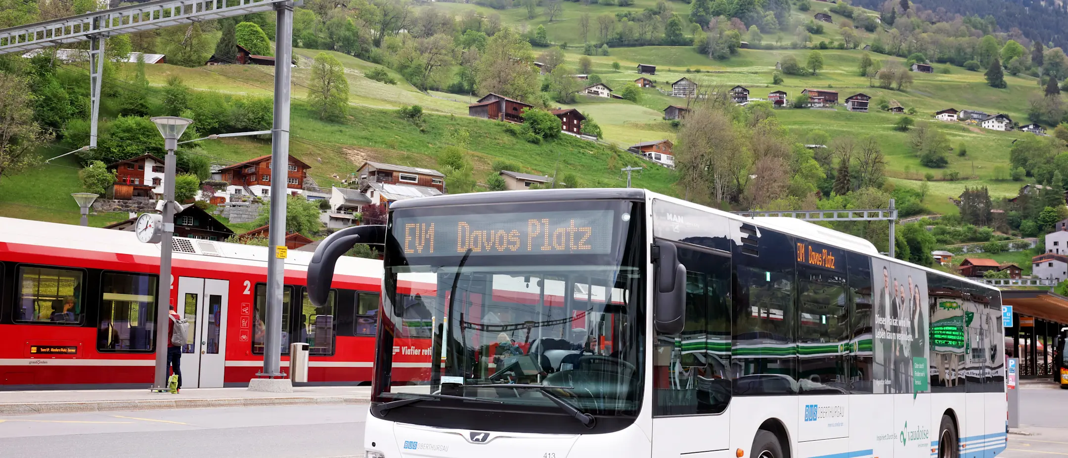 Ein hellblauer Bahnersatzbus mit dem Fahrtziel Davos Platz. Er steht in Küblis und im Hintergrund ist eine RhB-Bahn zu sehen.