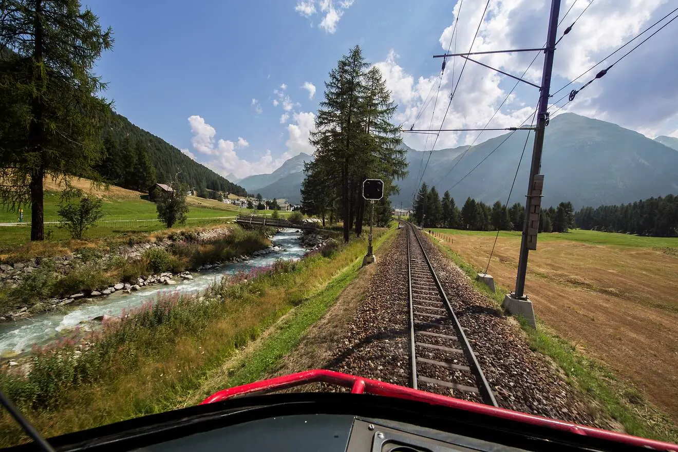 Führerstand einer Schmalspurbahn auf ein Gleis, das sich durch eine weite, grüne Landschaft mit Fluss und Bergen schlängelt.
