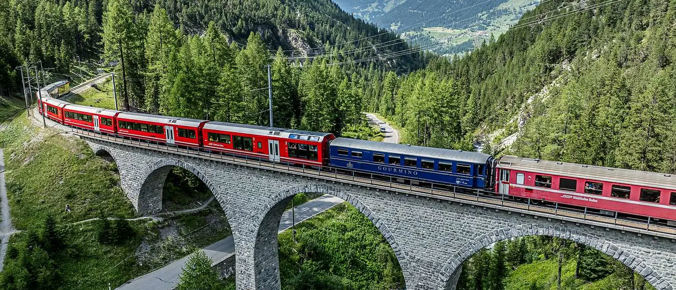 Der Speisewagen Gourmino als Teil eines Zugs der RhB überquert ein Viadukt inmitten einer alpinen Landschaft mit Wäldern.