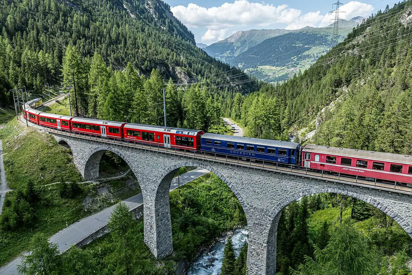 Der Speisewagen Gourmino als Teil eines Zugs der RhB überquert ein Viadukt inmitten einer alpinen Landschaft mit Wäldern.