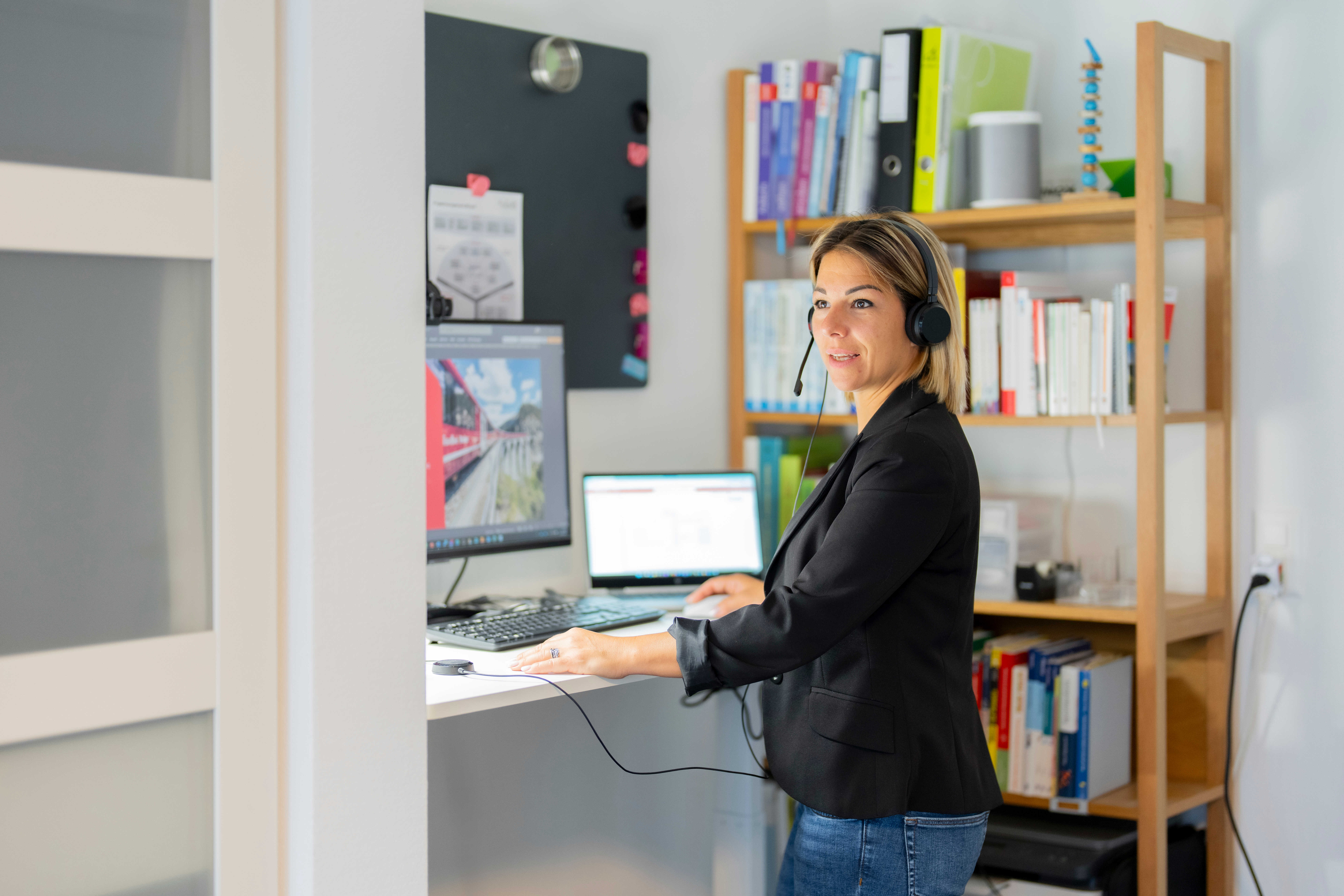 An employee is working on a desktop computer and a laptop, wearing headphones, in her home office