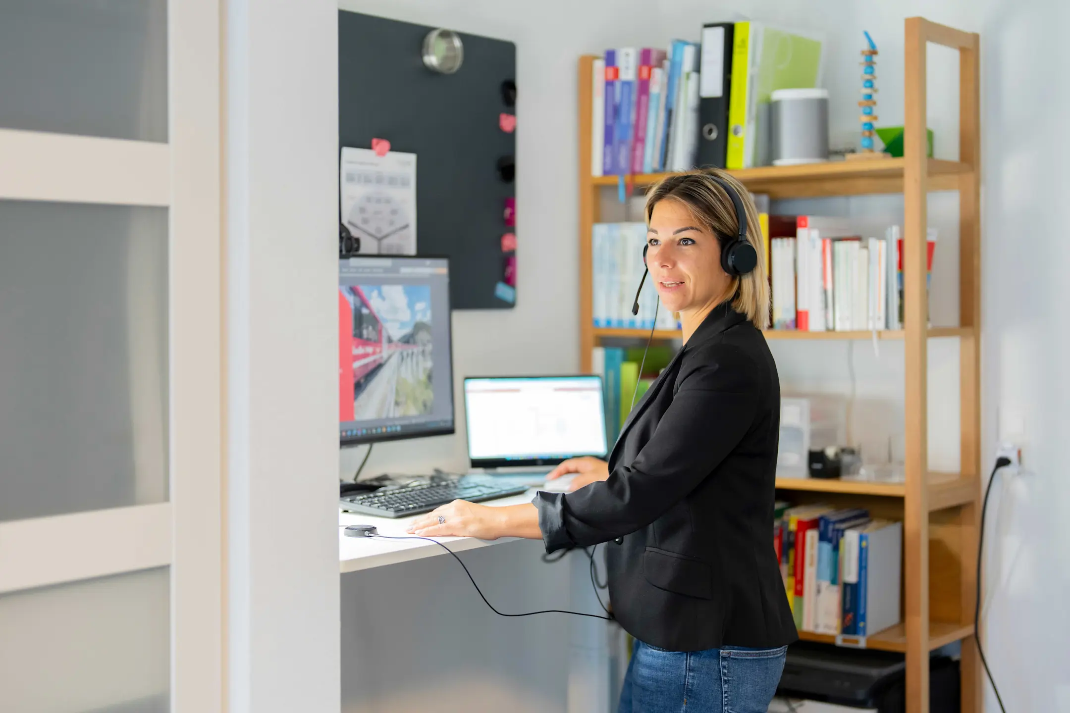 An employee is working on a desktop computer and a laptop, wearing headphones, in her home office