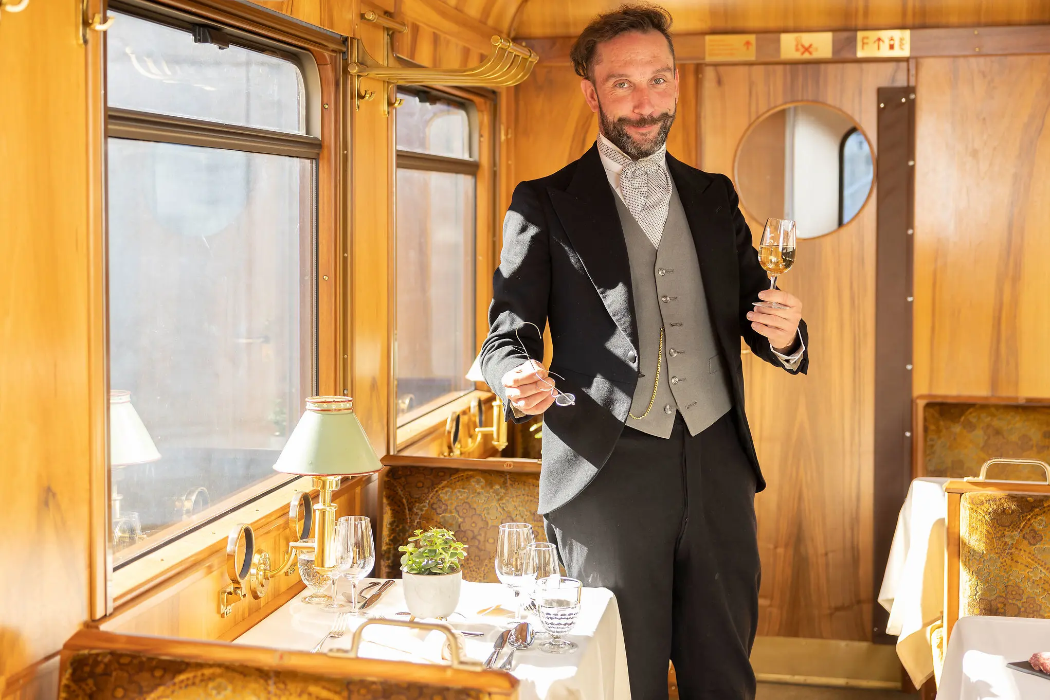 Ein Mann im historischen Frack mit Monokel und Champagnerglas steht in einem nostalgisch eingerichteten Speisewagen.