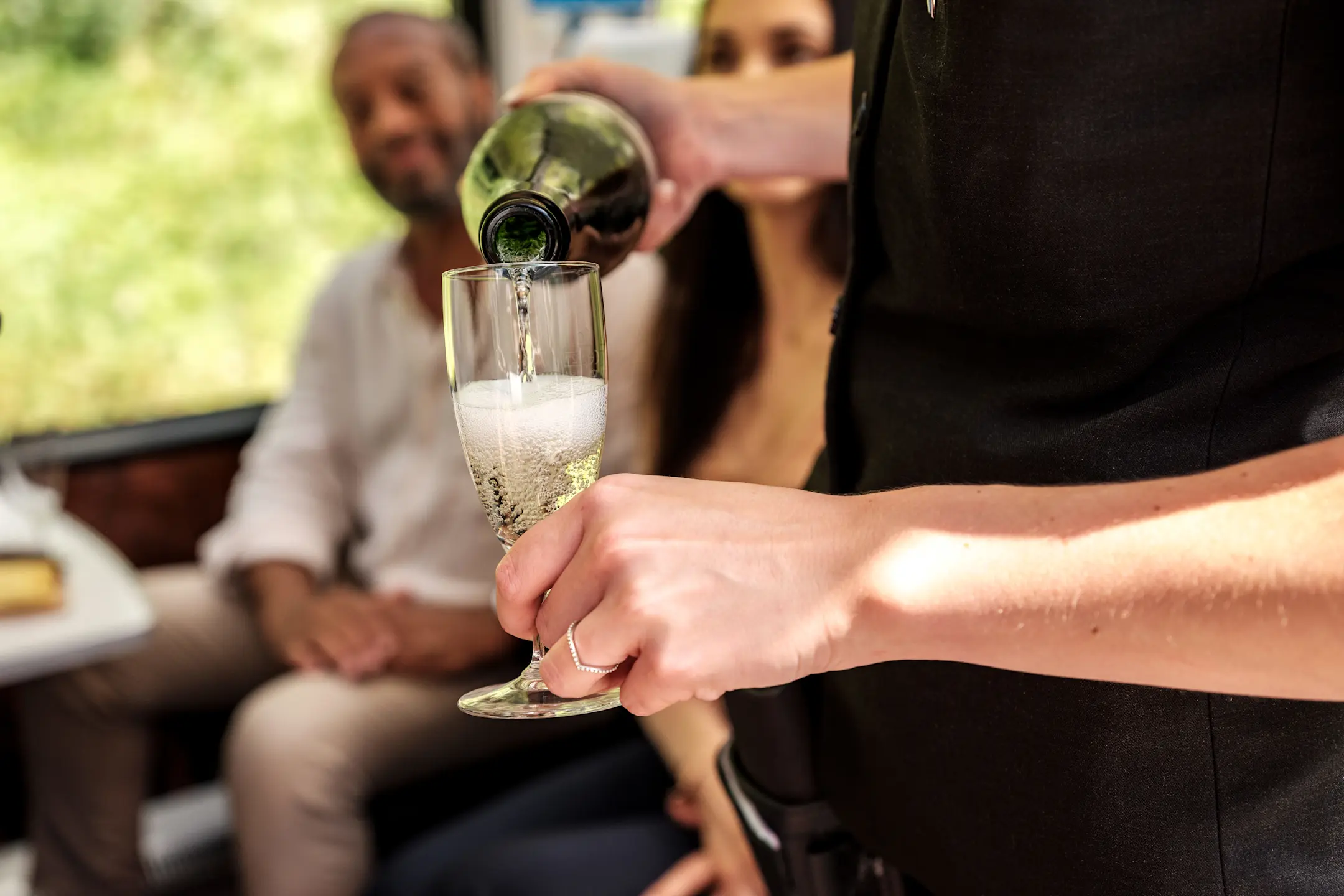 Ein Zugmitarbeiter schenkt Sekt in ein Glas ein, während zwei Fahrgäste entspannt im Hintergrund sitzen.