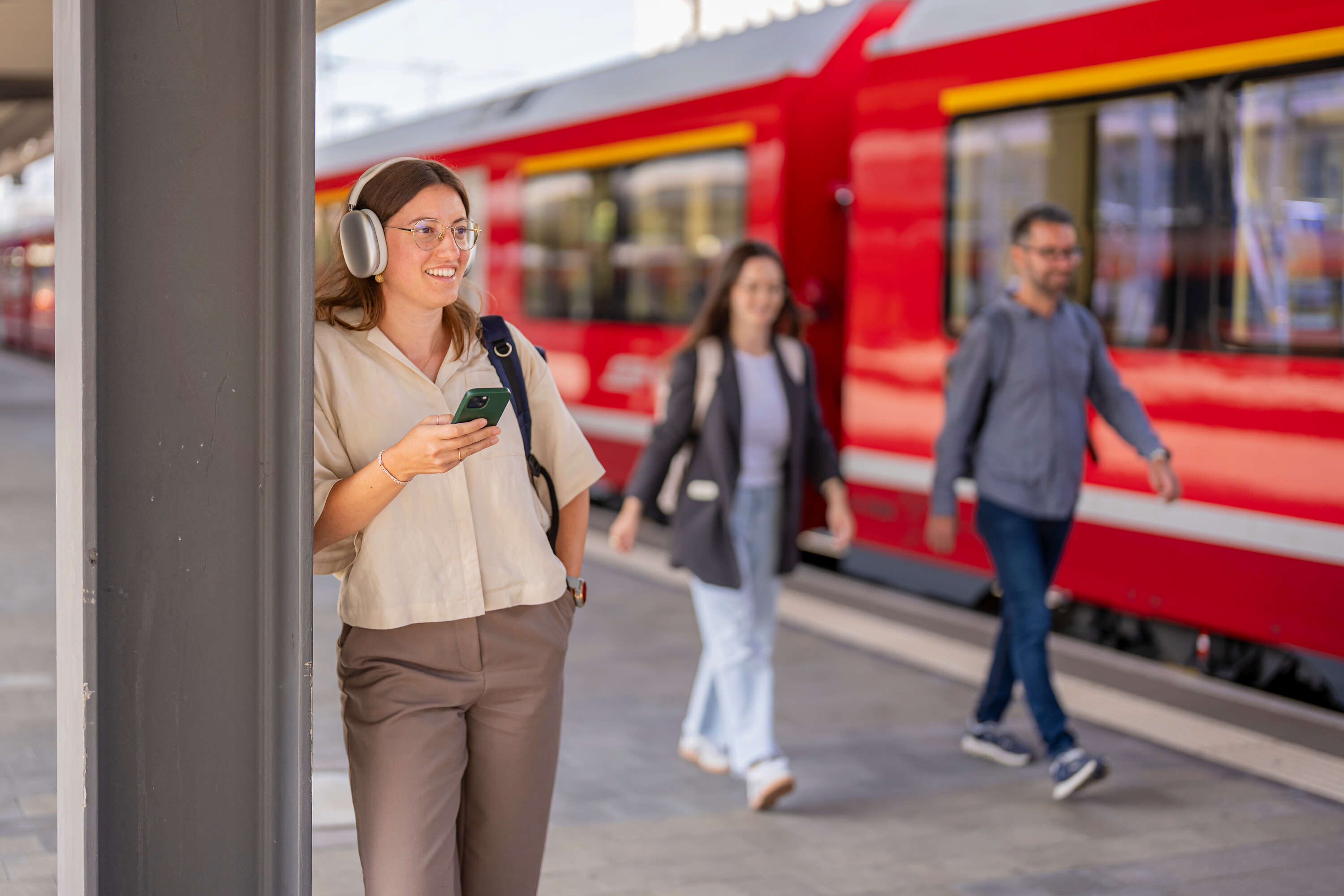 Eine junge Frau mit Smartphone und Kopfhörern auf einem Bahnsteig vor einem Zug und zwei passierenden Menschen.