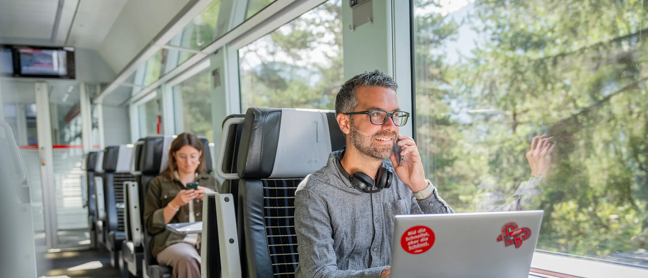 Ein Mann sitzt mit seinem Laptop an einem Tisch im Zug und telefoniert.
