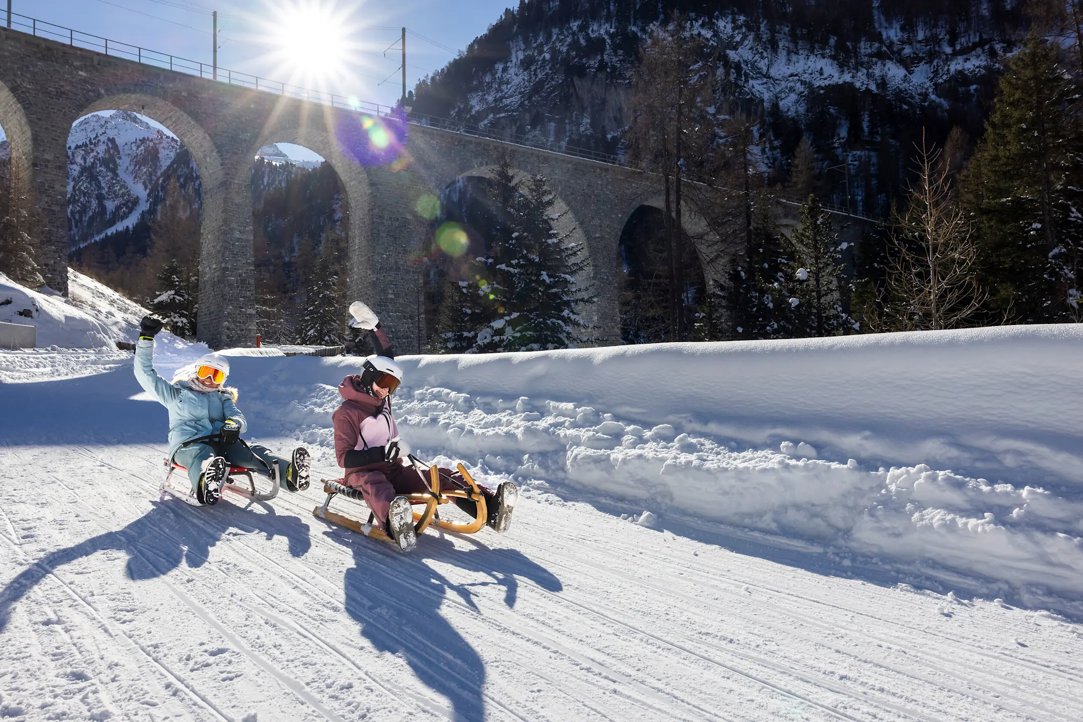 Mehrere Personen rodeln auf einer präparierten Schneepiste unter einem Viadukt hindurch, über den ein roter Zug fährt.